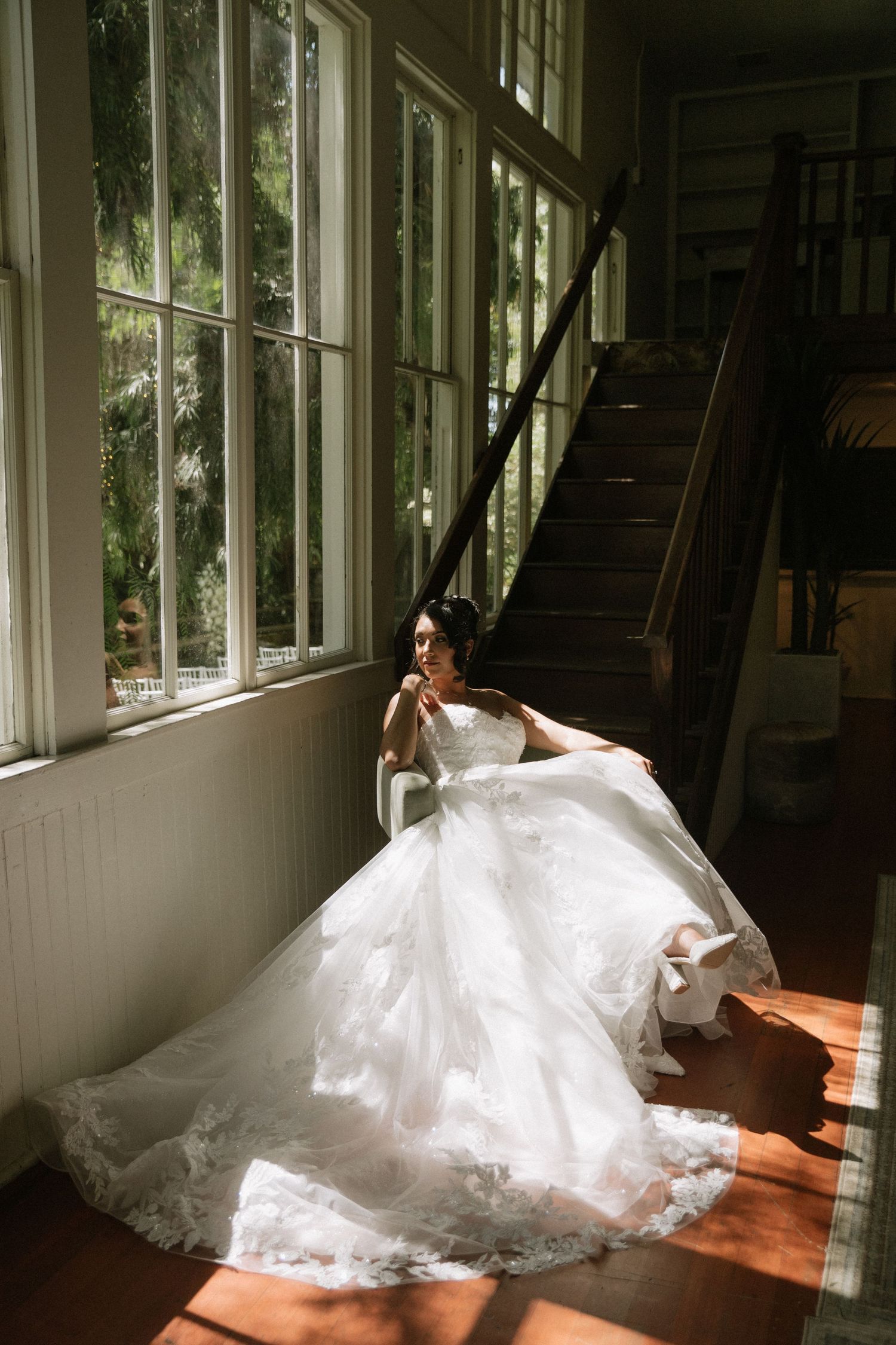 Bride in flowing wedding gown poses by sunlit windows in vintage interior.