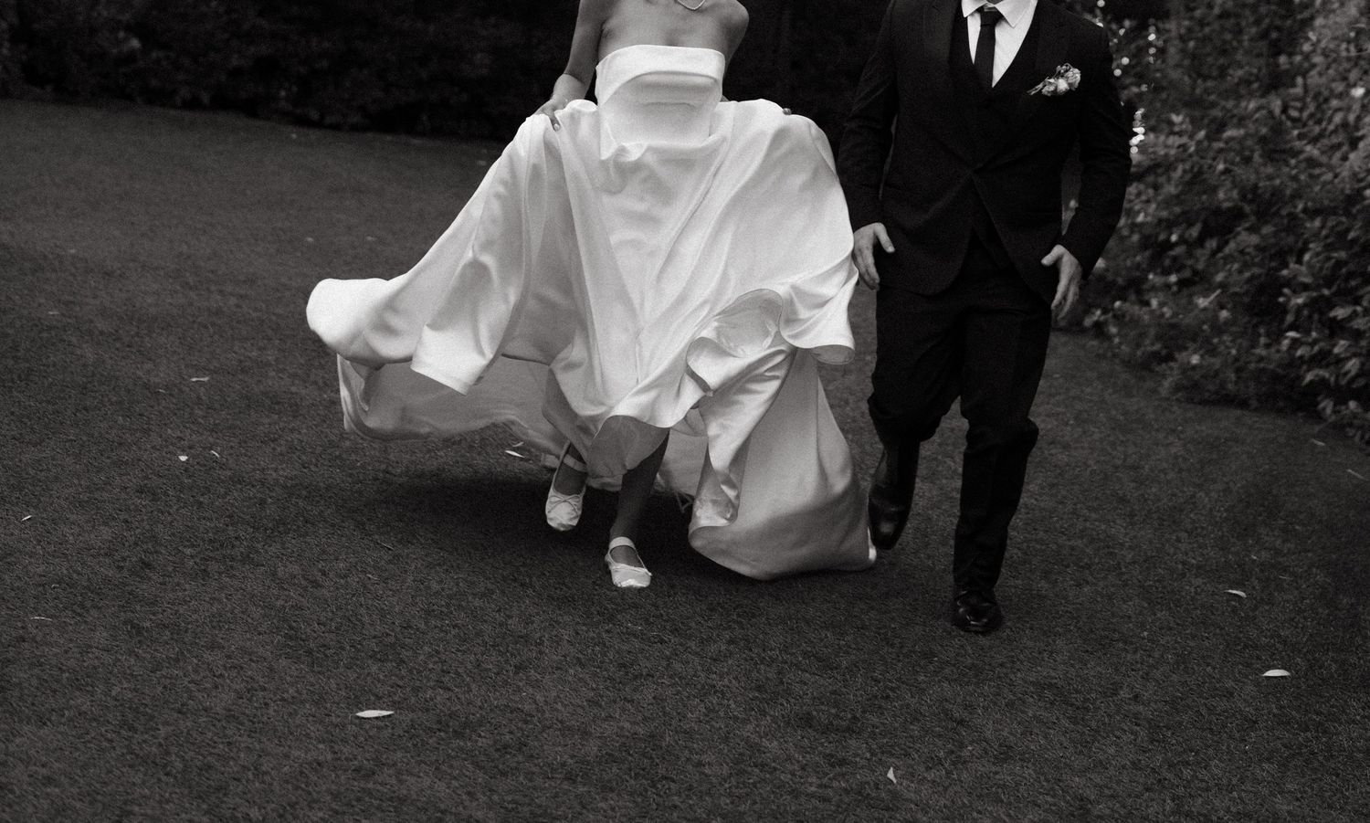 Black and white photo showing the bottom of a flowing white wedding dress and dress shoes walking with a suit.