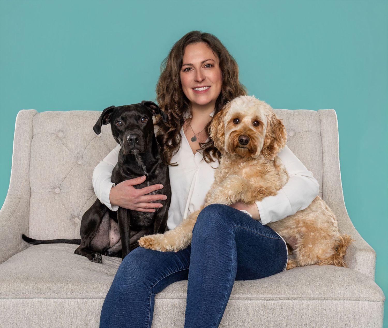 Woman smiling on gray sofa with black Labrador mix and golden Doodle dogs against teal background.