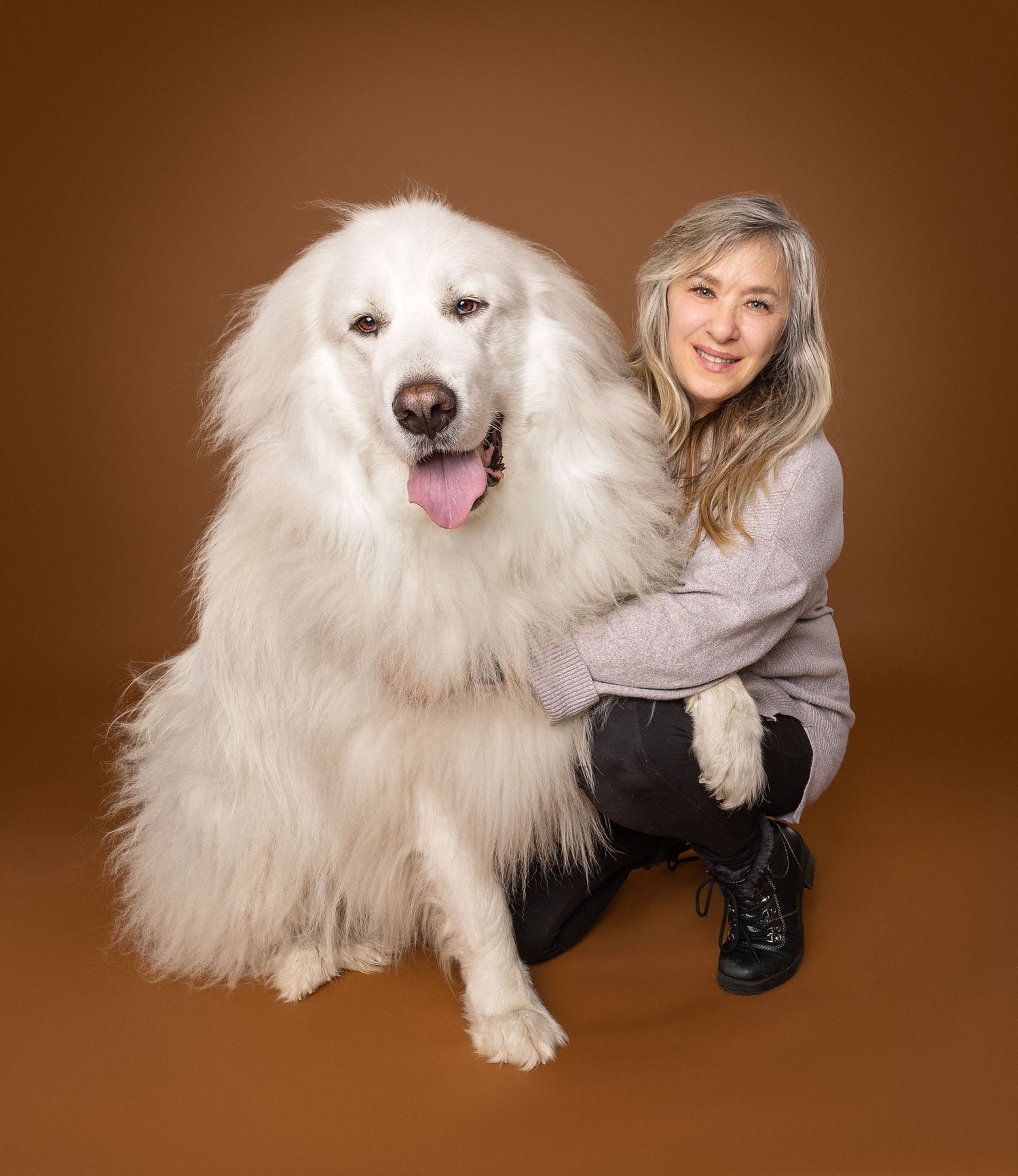 Woman smiling beside a large fluffy white Great Pyrenees dog on a brown studio background.