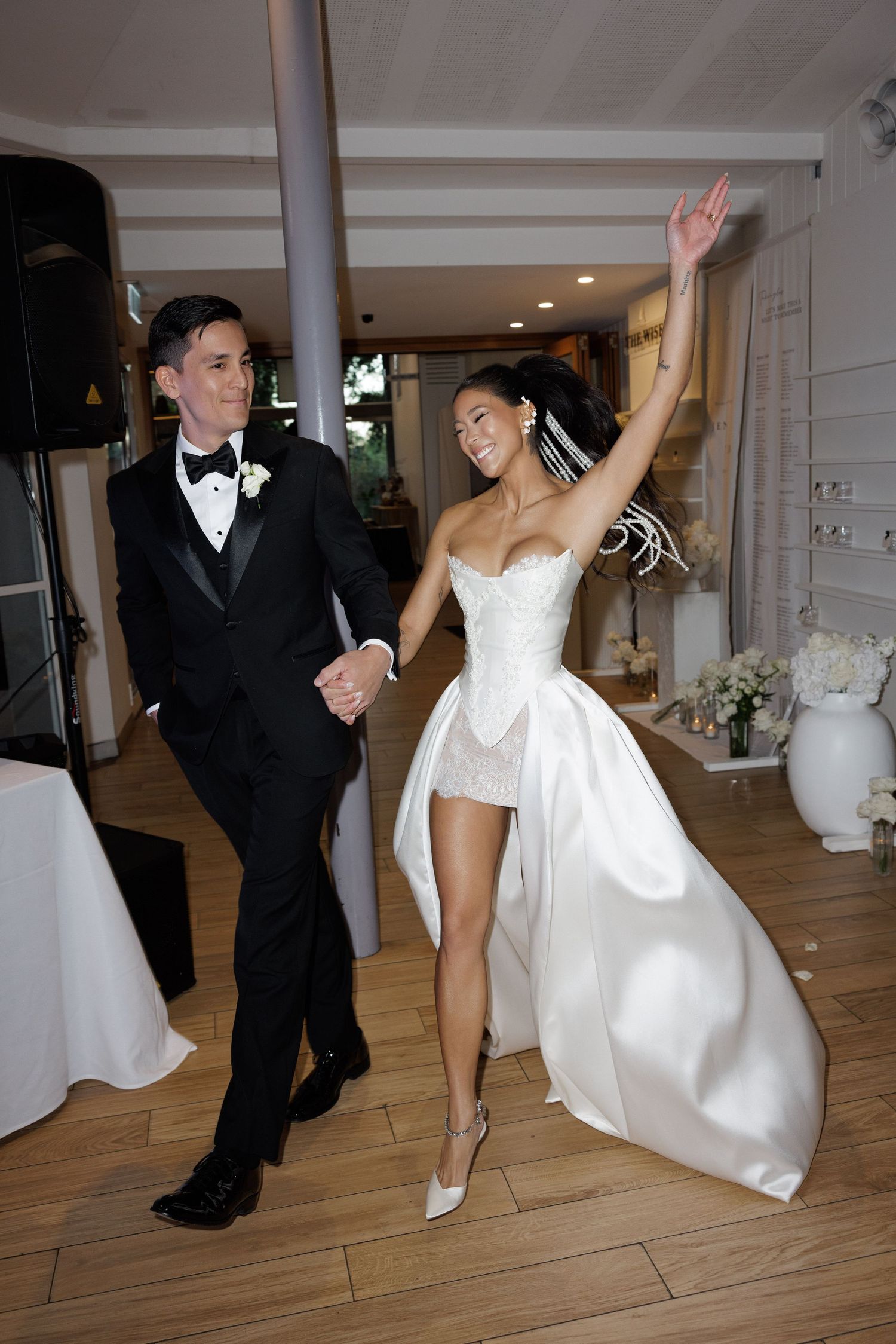 Bride in strapless mini wedding dress waves joyfully while entering reception with groom.