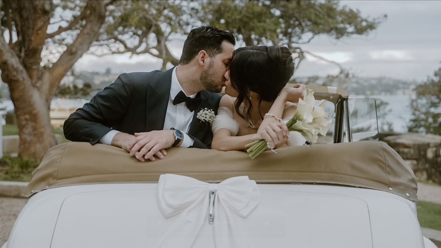 Bride and groom sharing a kiss in a vintage convertible car with scenic waterfront backdrop.