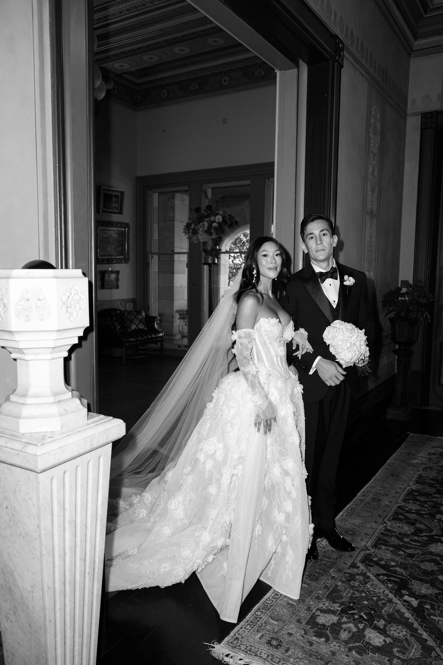 Black and white photo of bride in floral gown with cathedral veil walking with groom in tuxedo.