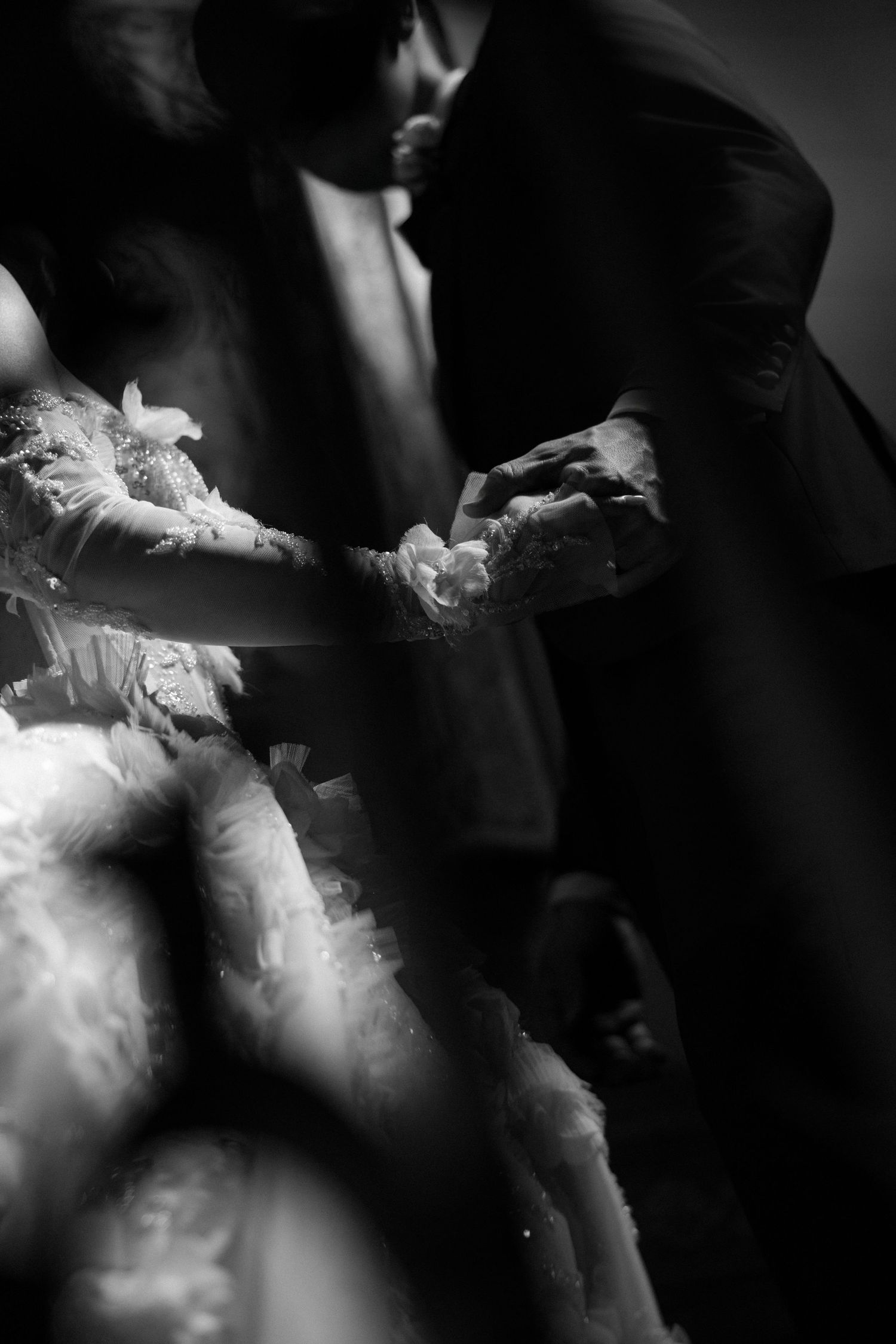 Black and white close-up of groom holding brides lace-gloved hand in soft dramatic light.