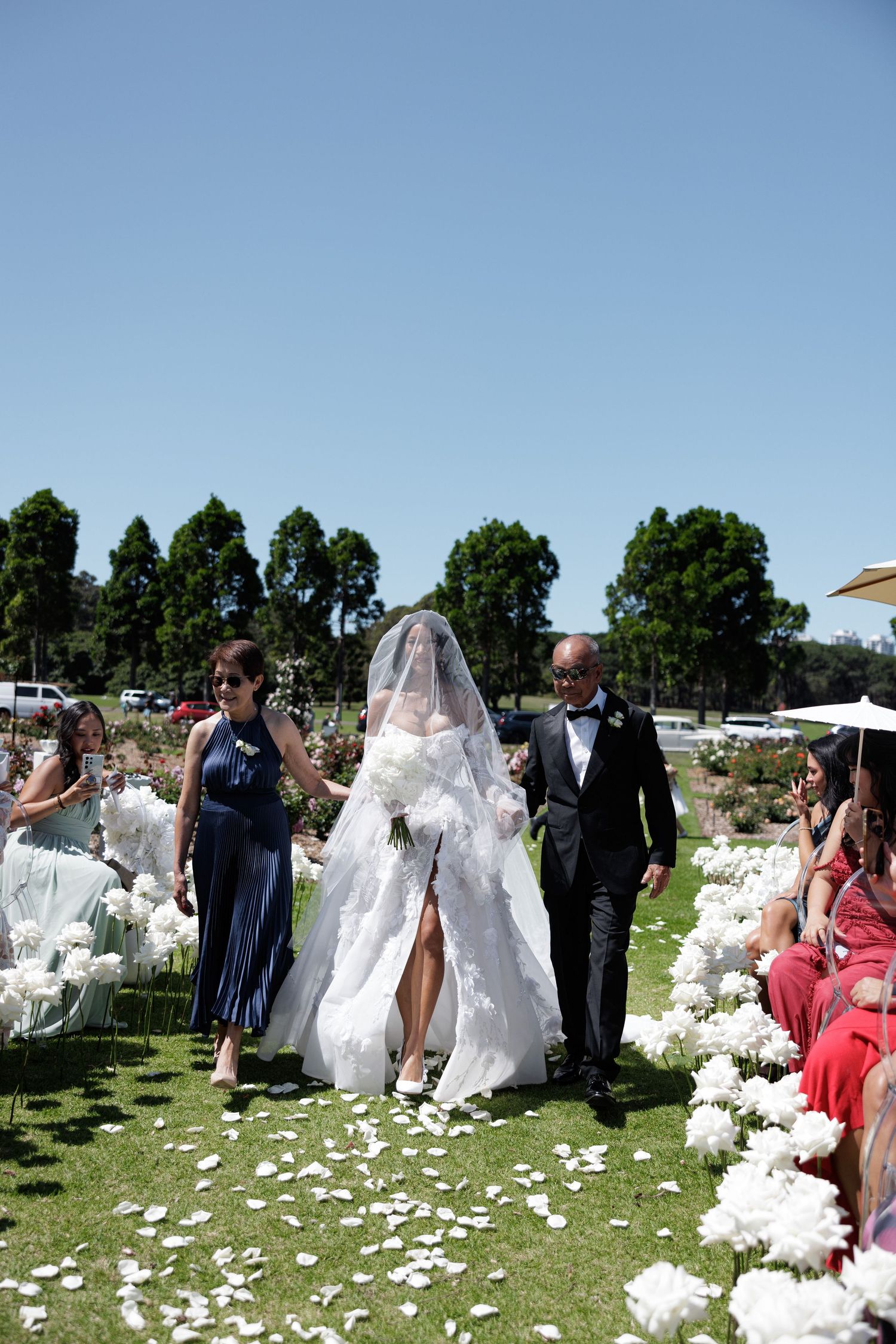 Bride in stunning lace wedding gown with veil walking down outdoor aisle flanked by white floral arrangements.