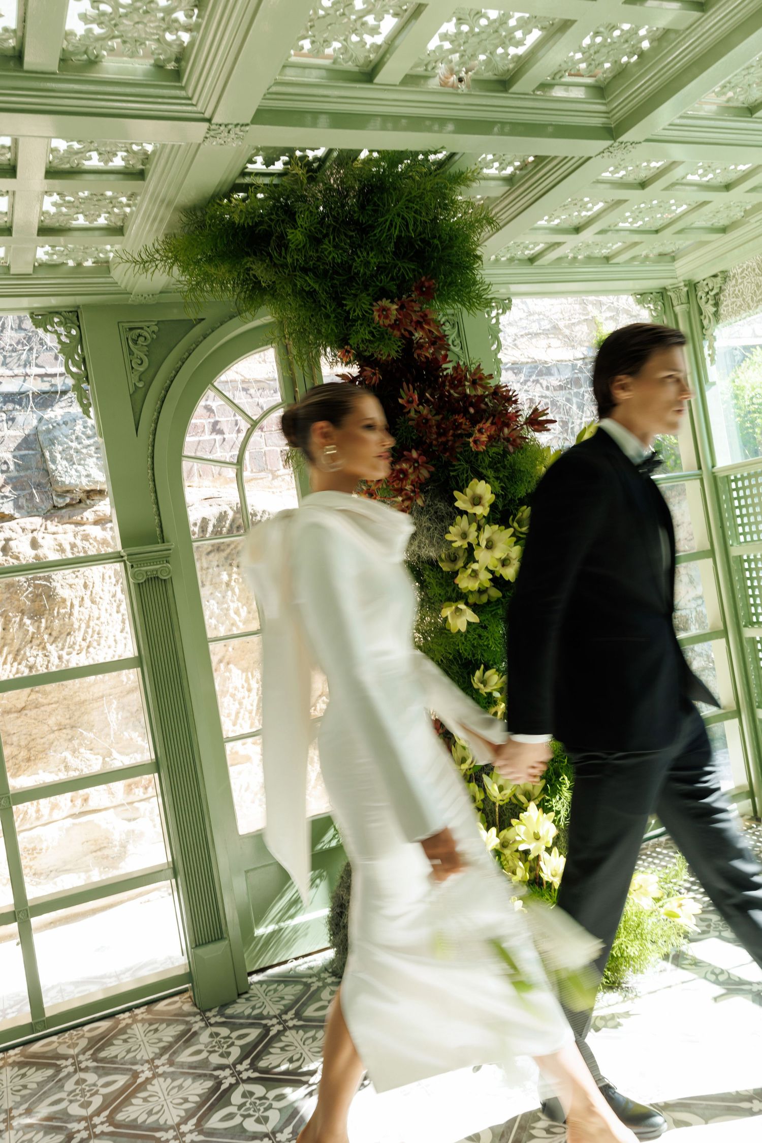 Bride and groom walk hand-in-hand through a flower-adorned greenhouse corridor in a dreamy motion-blur wedding photo.