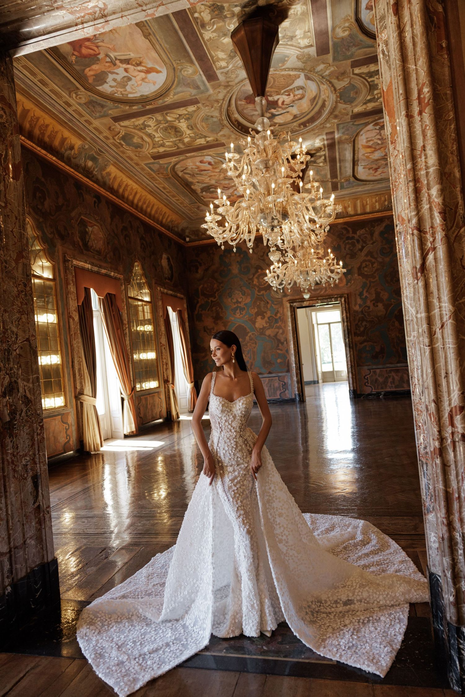 Bride in ornate lace mermaid gown with long train stands in opulent baroque palace hall with chandelier.