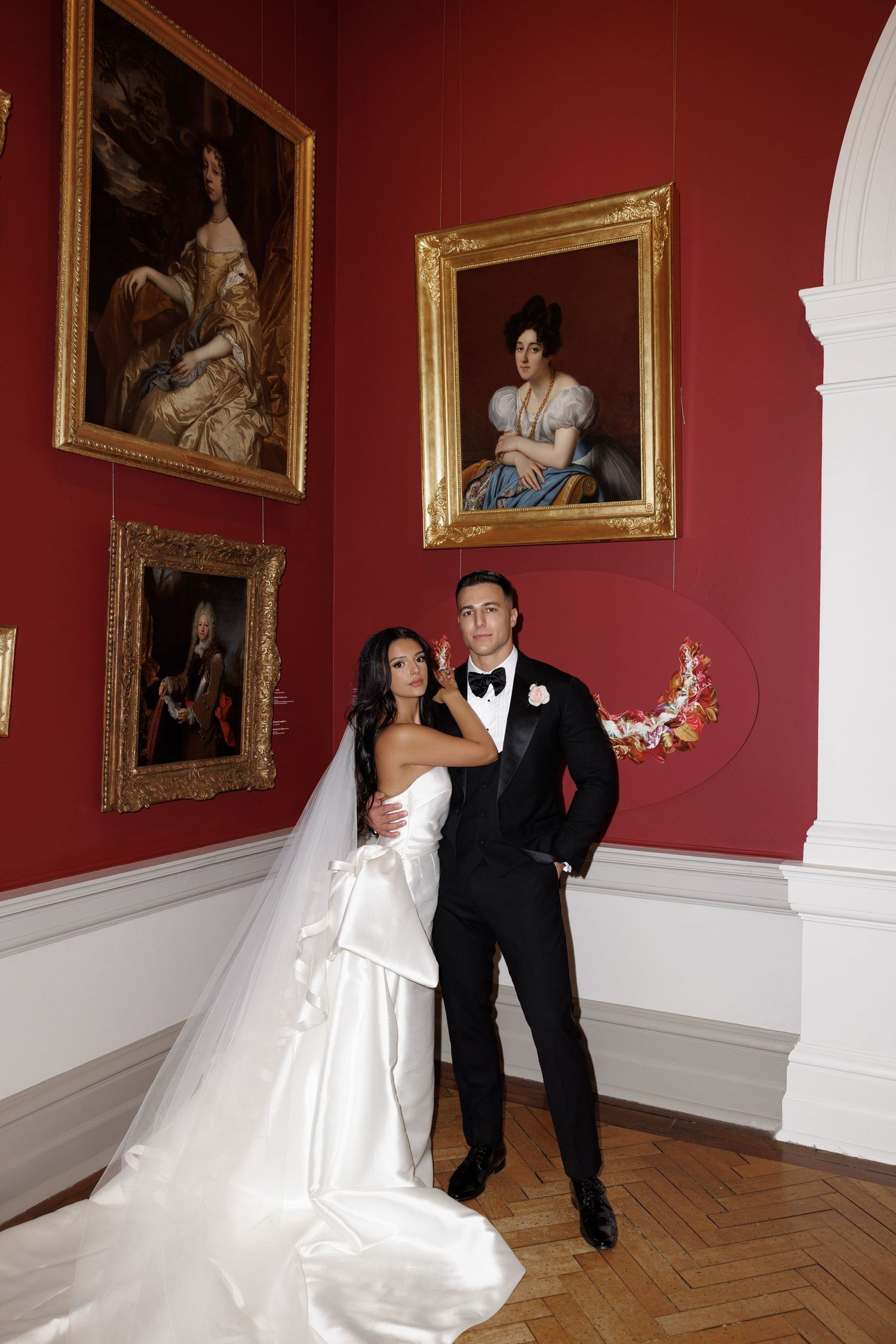 Bride and groom pose elegantly in art gallery with classic portrait paintings on red walls.