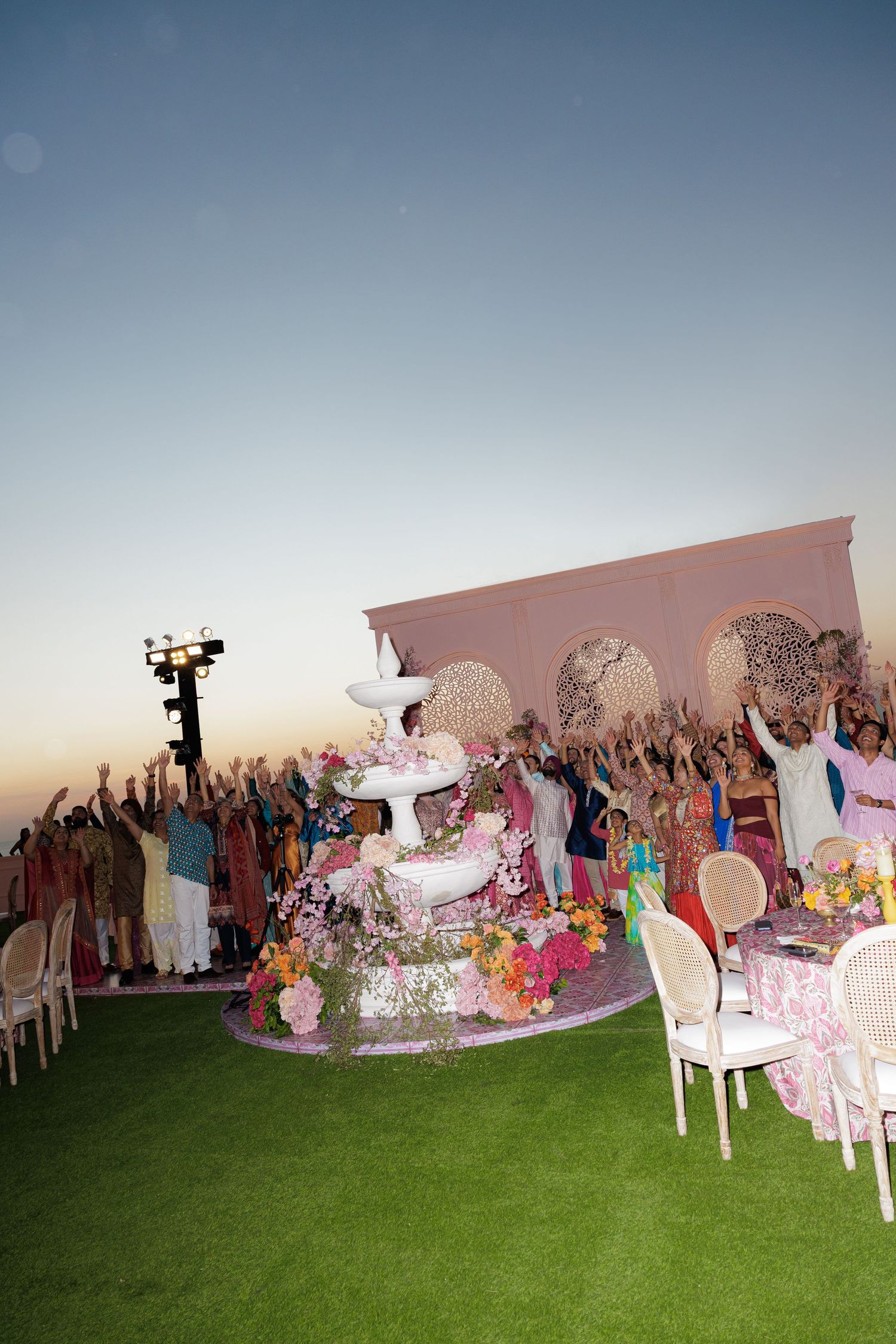 Outdoor rooftop celebration at sunset with guests raising hands near a white fountain and floral decorations.