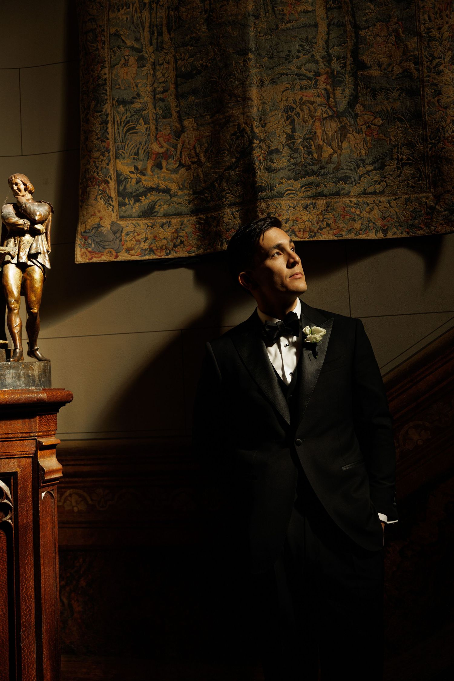 Elegant groom in black tuxedo leans against wall beside bronze statue and ornate tapestry in dramatic light.