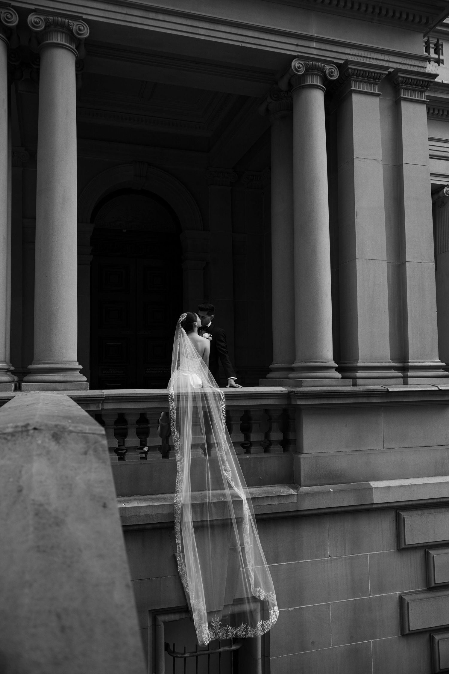 Bride with long lace veil standing on grand building balcony with tall columns, black and white.