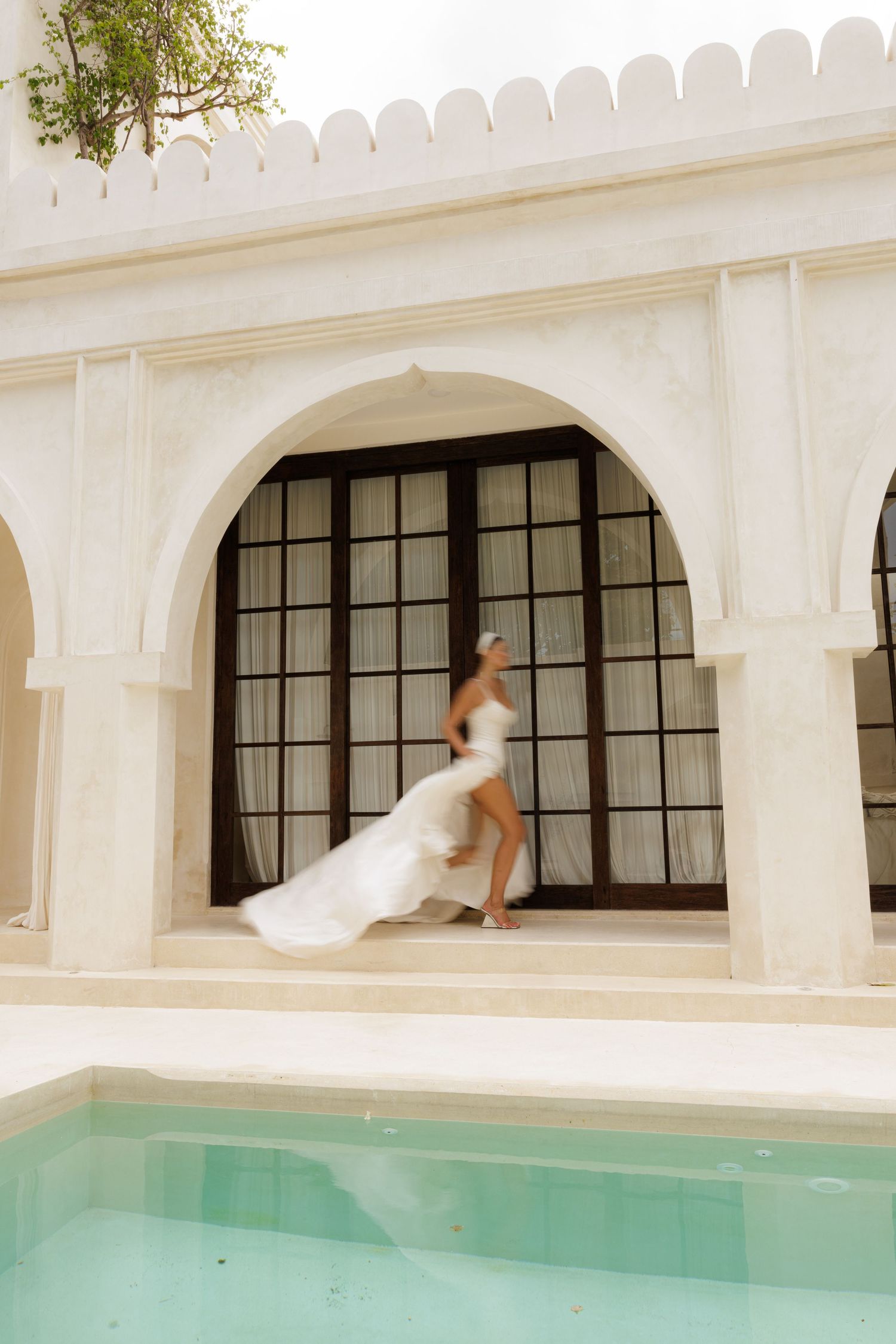 Bride in flowing white wedding dress walking through elegant arched colonnade beside turquoise pool.