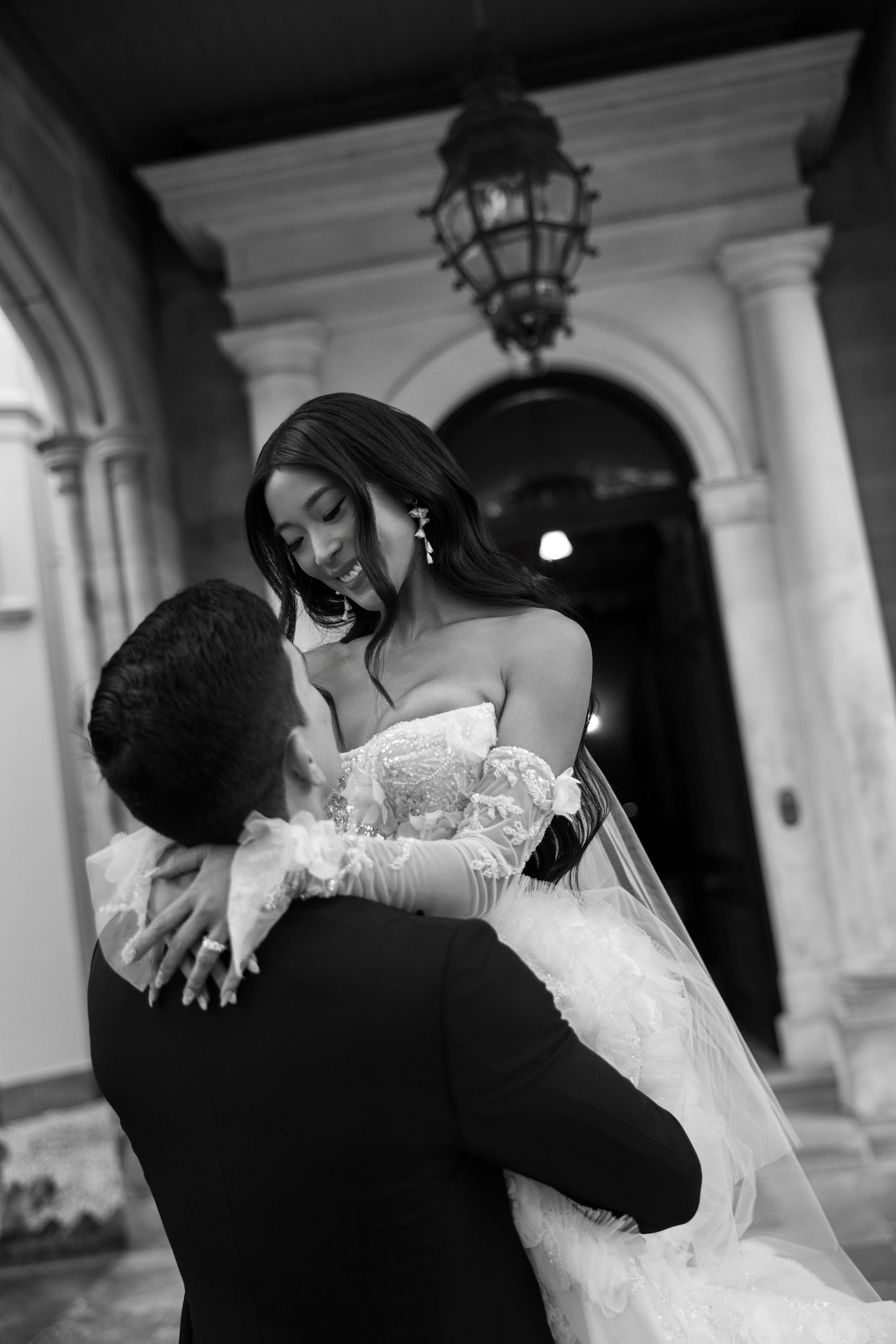 Black and white photo of groom lifting bride in wedding dress under ornate archway.