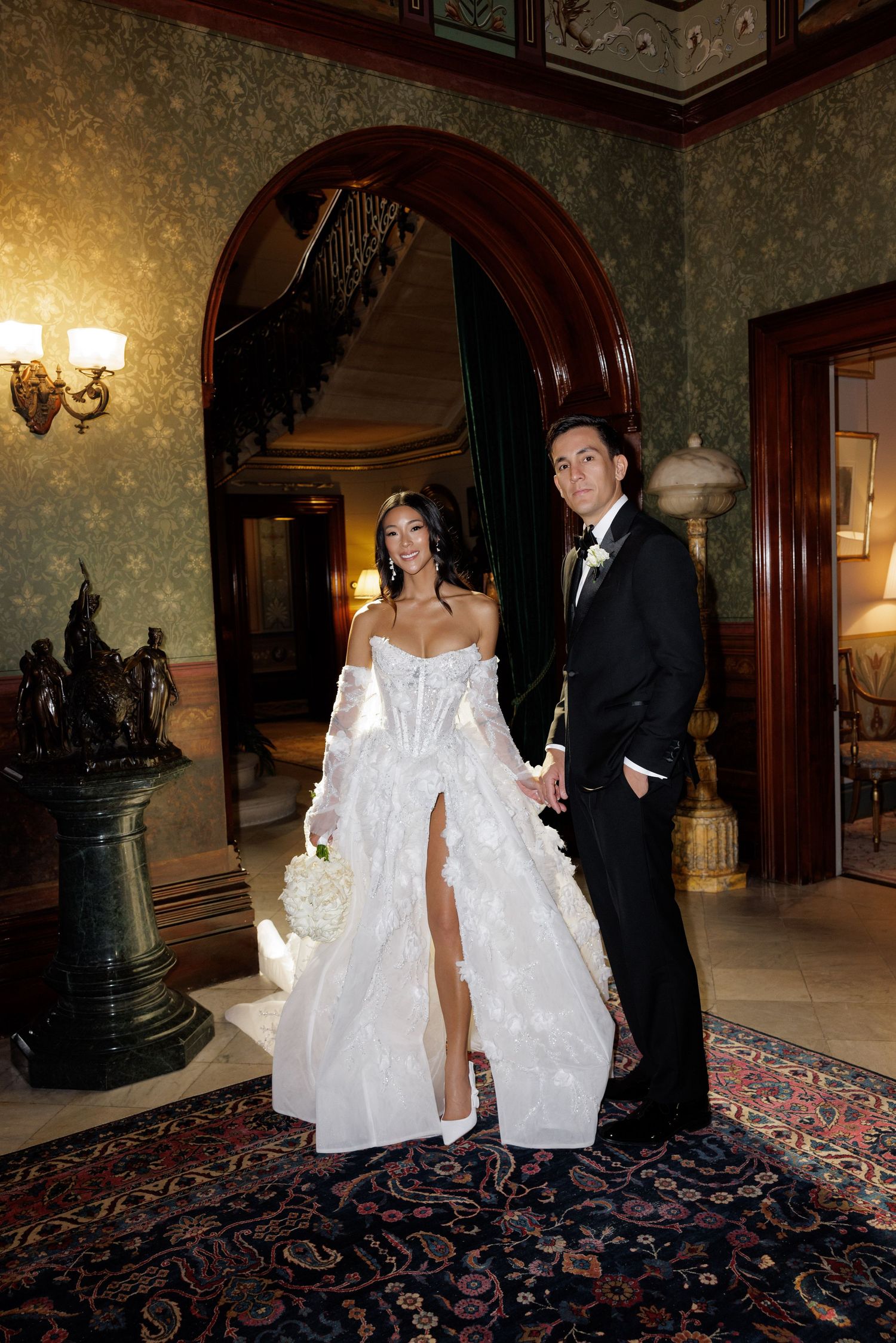 Bride in strapless white corset gown with high slit and groom in black tuxedo posing in ornate mansion foyer.