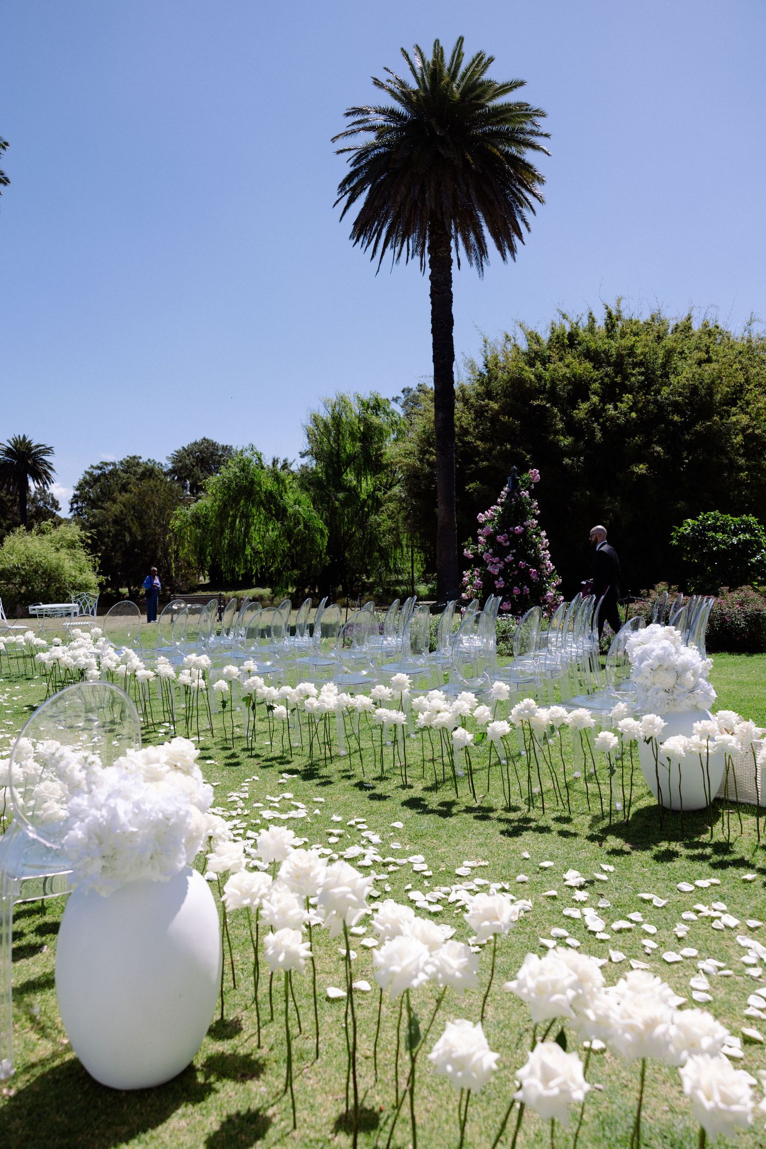 Elegant outdoor wedding ceremony setup with white floral arrangements and ghost chairs on green lawn with palm trees.