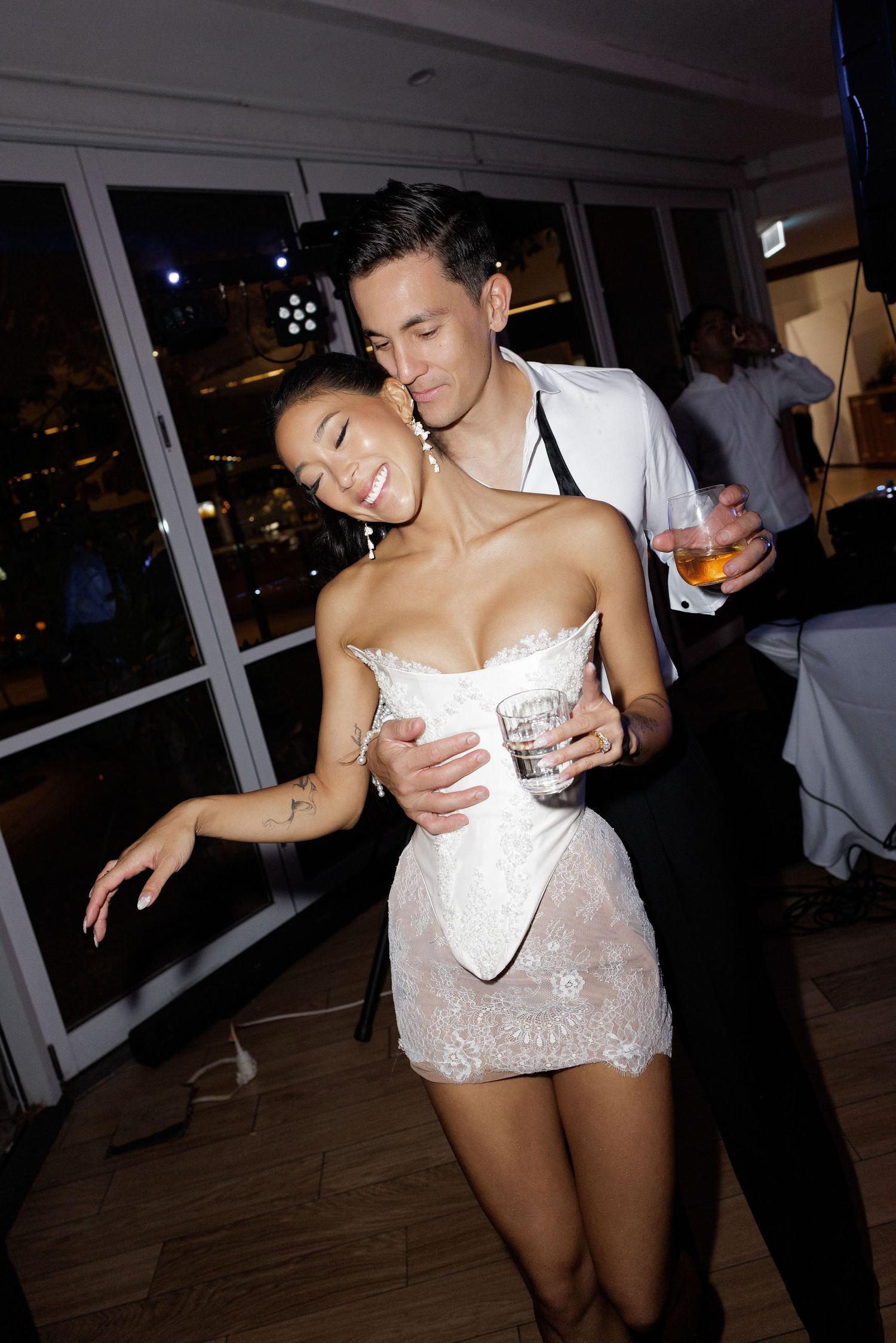 Bride in white lace mini dress dances with groom holding cocktail at wedding reception.