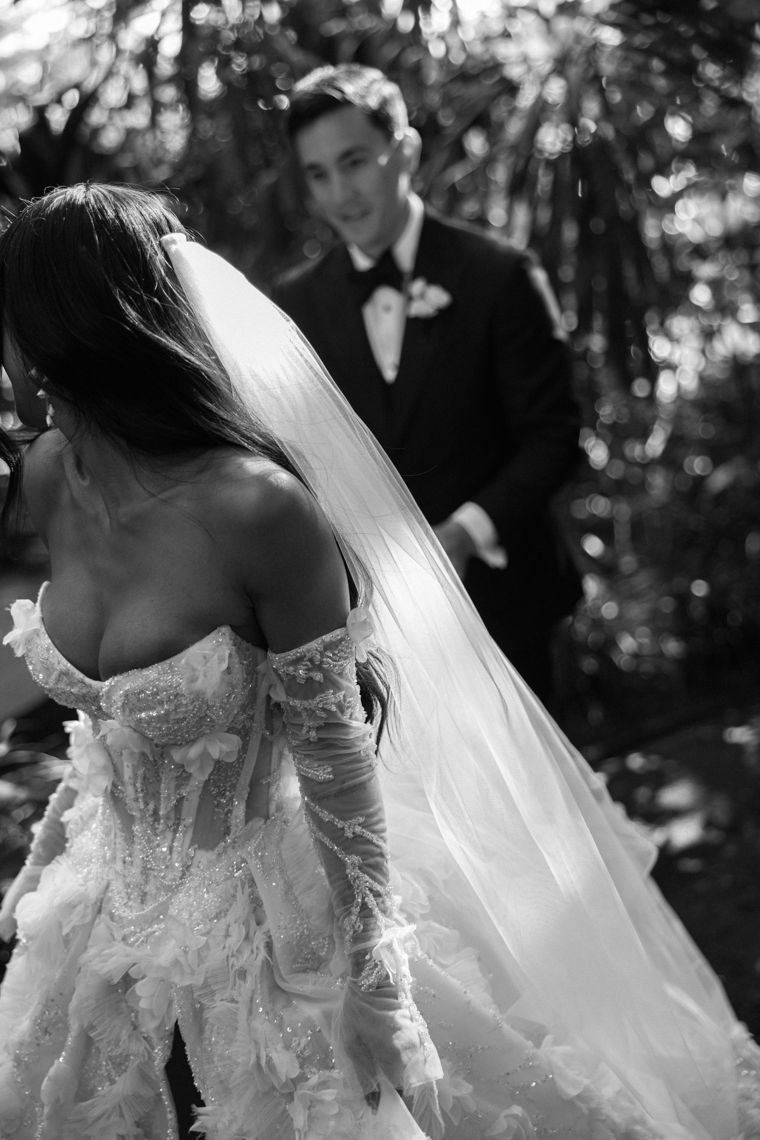Black and white photo of bride in lace gown with veil approaching groom outdoors.