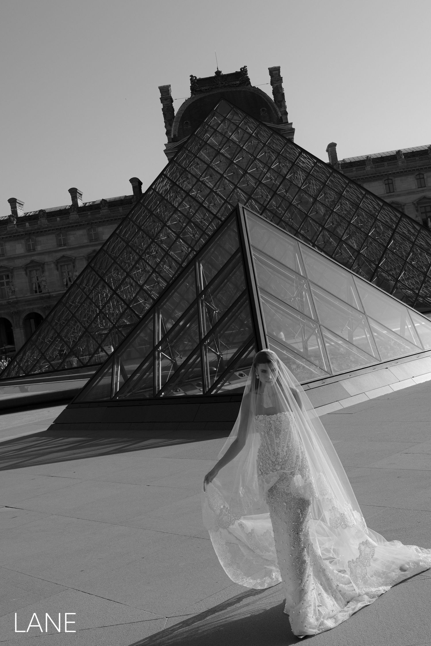 Bride in lace wedding dress and veil poses before the Louvre Pyramid in Paris.