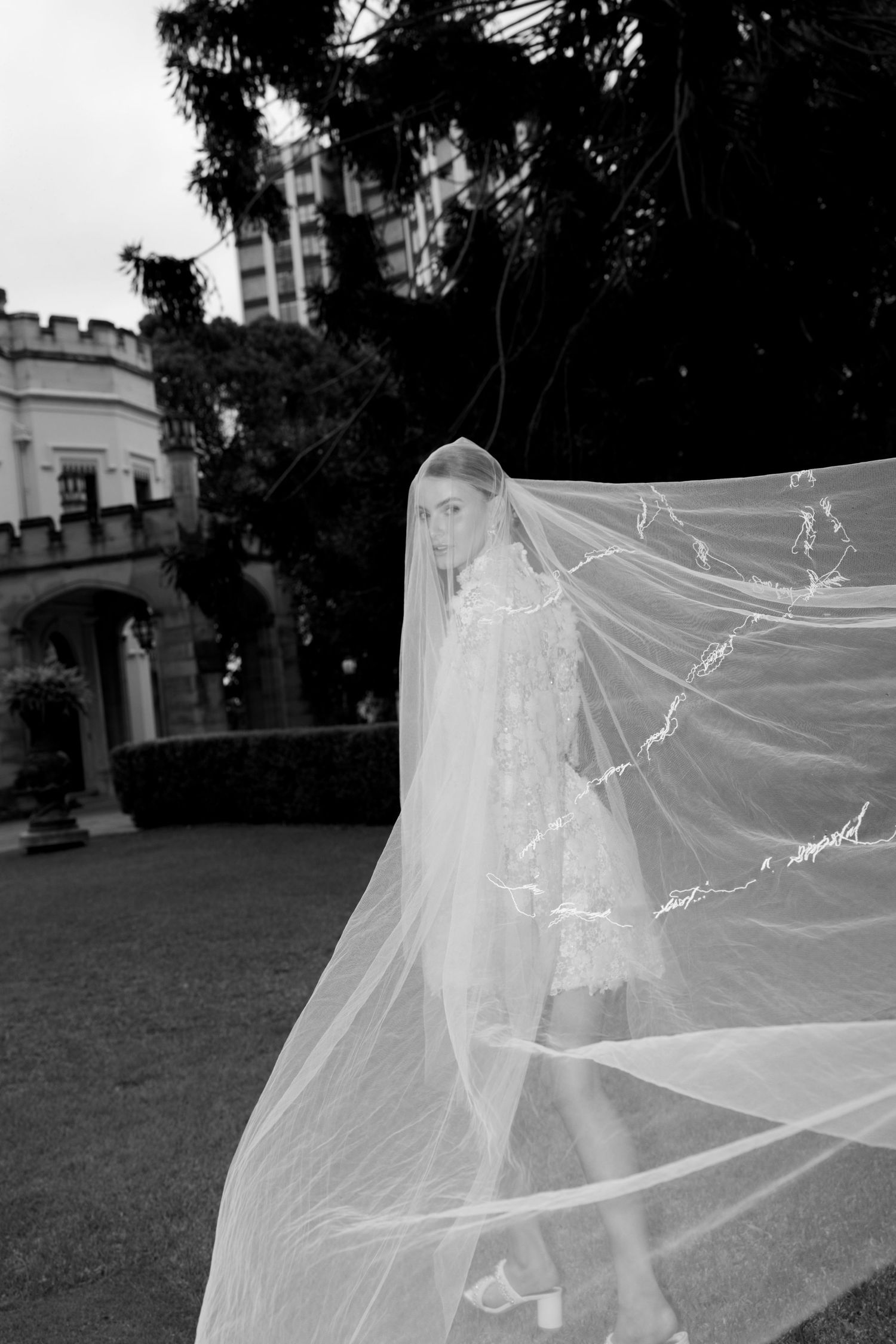 Bride in lace gown with flowing veil billowing in the wind outside a historic stone building.