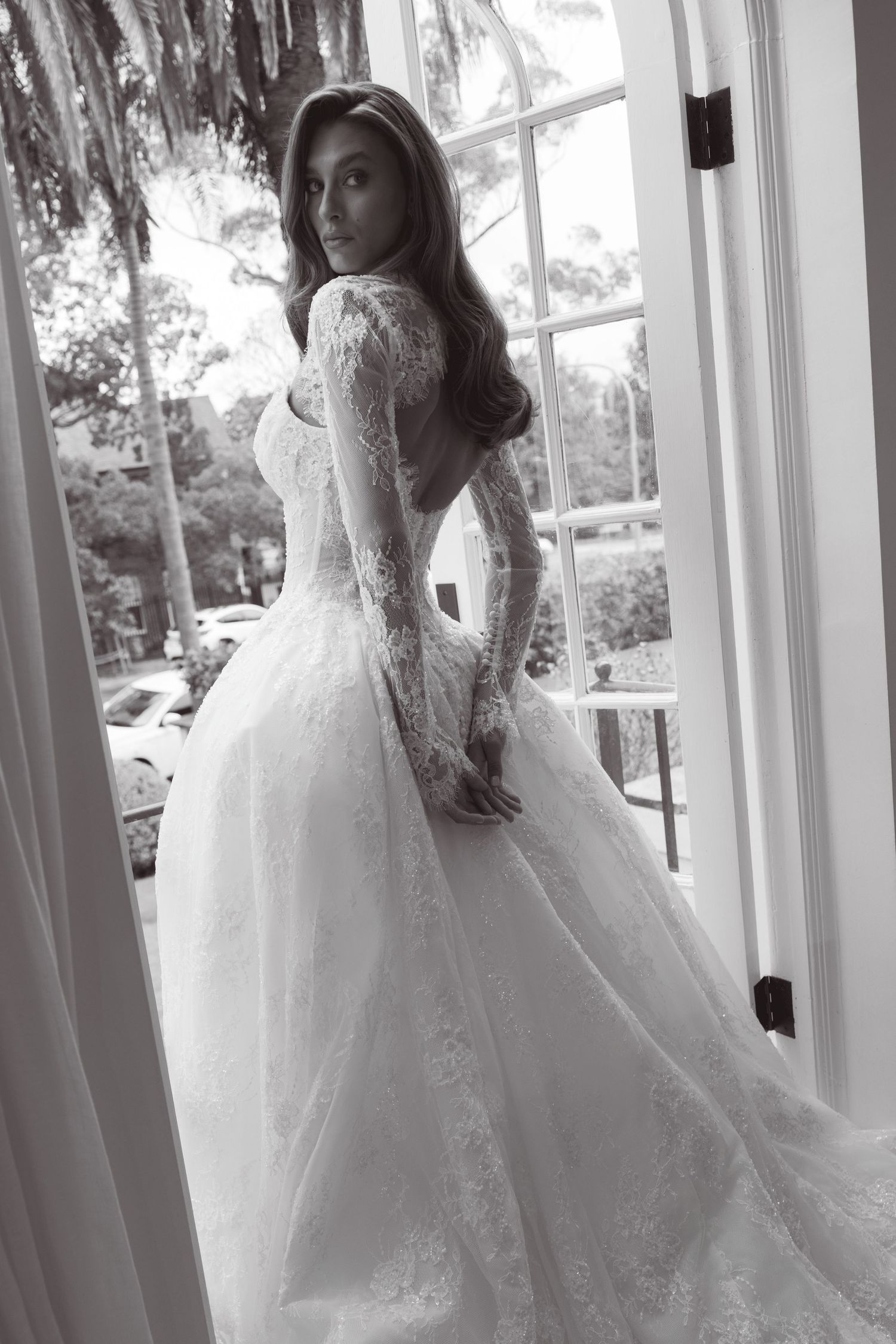 Elegant bride in a lace long-sleeve backless ball gown wedding dress, posing by French doors in black and white.