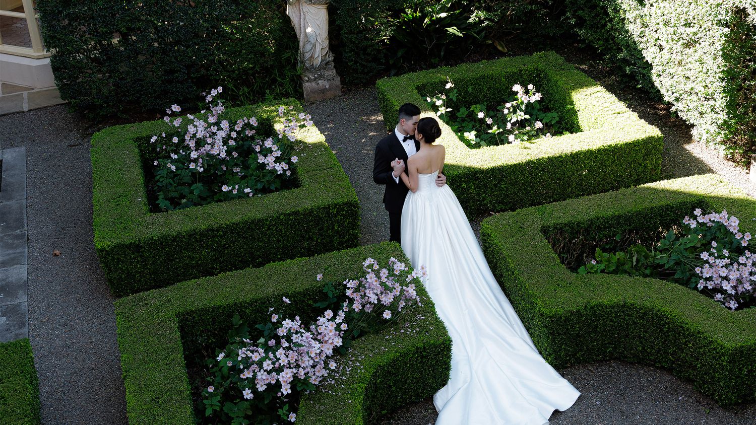 Aerial view of bride and groom embracing in formal garden with manicured hedges and pink flowers.