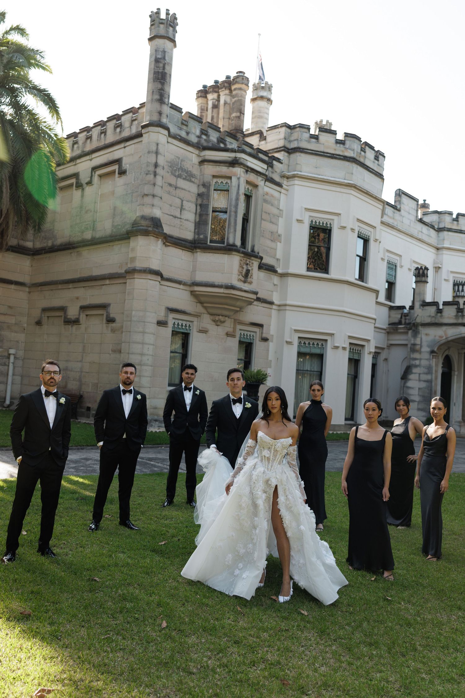Wedding party in black formal wear flanks bride and groom before Gothic castle venue on sunny day.
