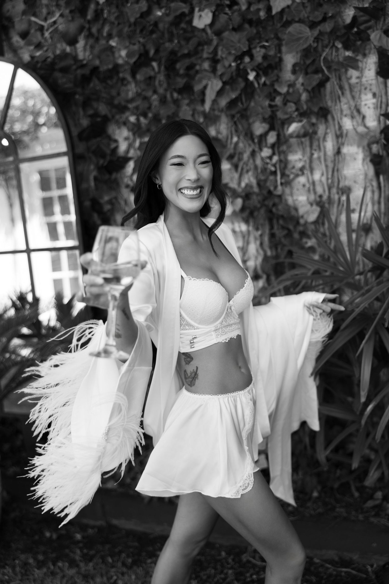 Smiling bride in white lingerie and feathered robe poses outdoors in black and white photo.