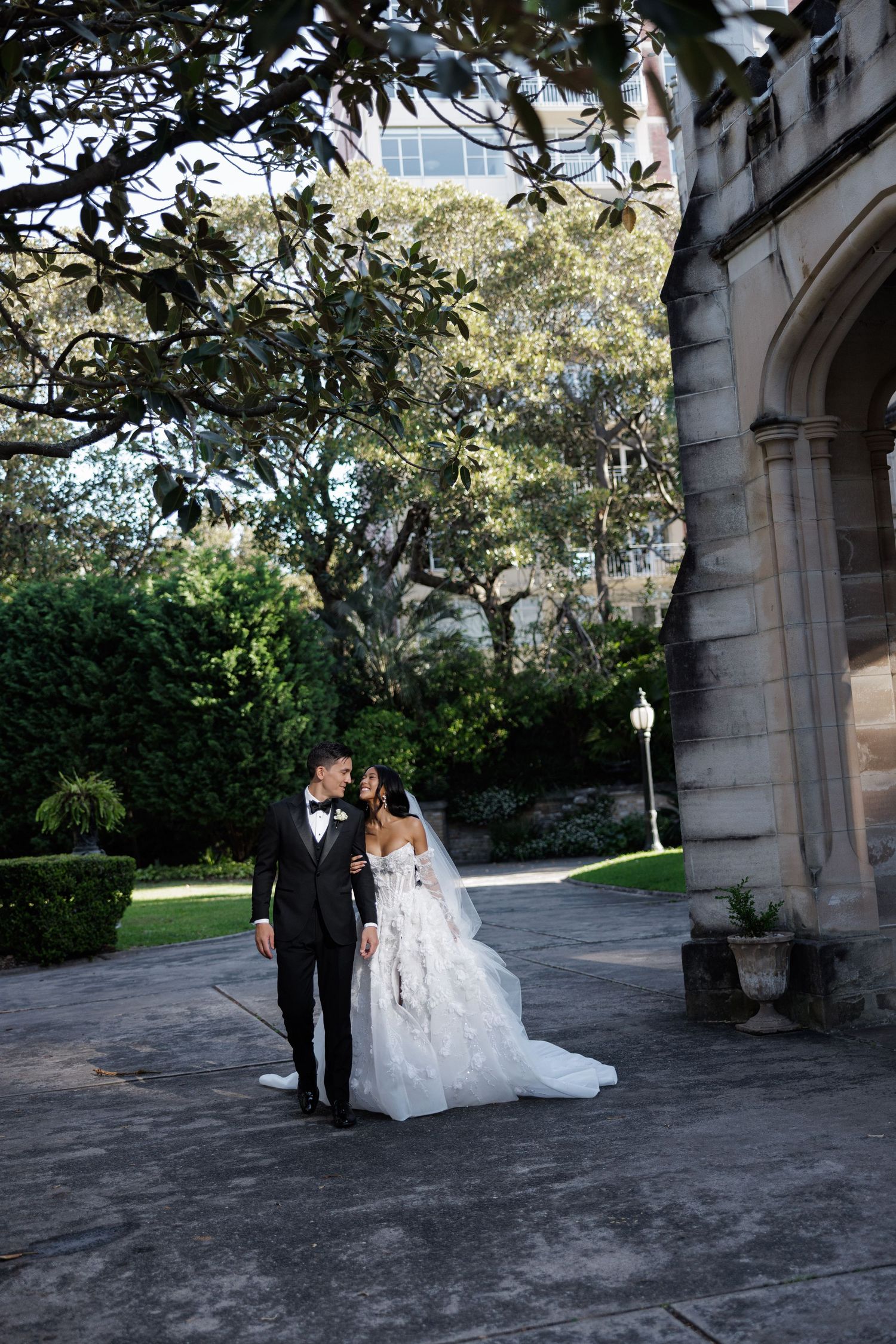 Bride and groom share tender moment beneath trees beside Gothic stone arch at elegant wedding venue.