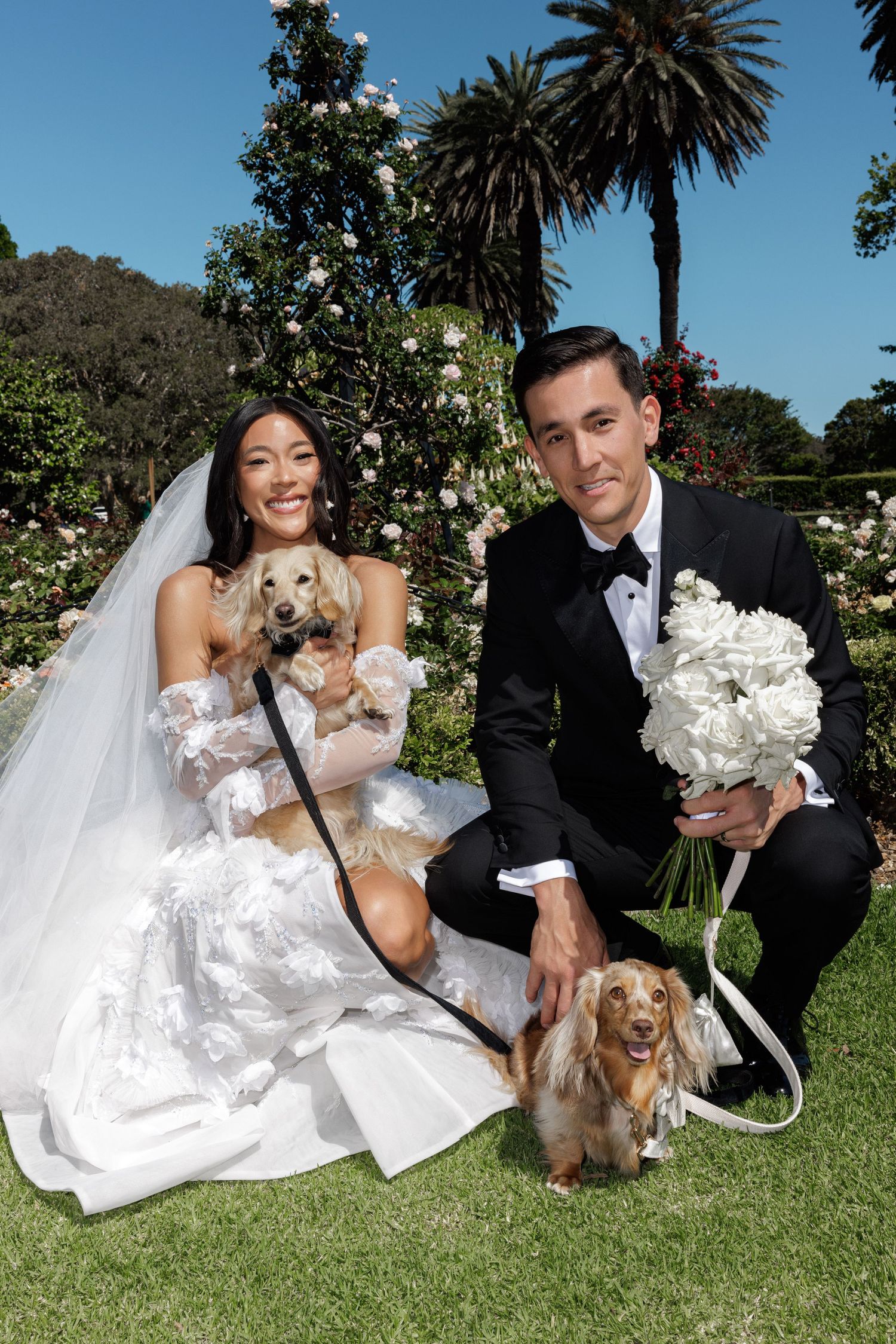 Newlyweds posing with two golden dachshunds in a lush garden, bride in white gown holding white floral bouquet.