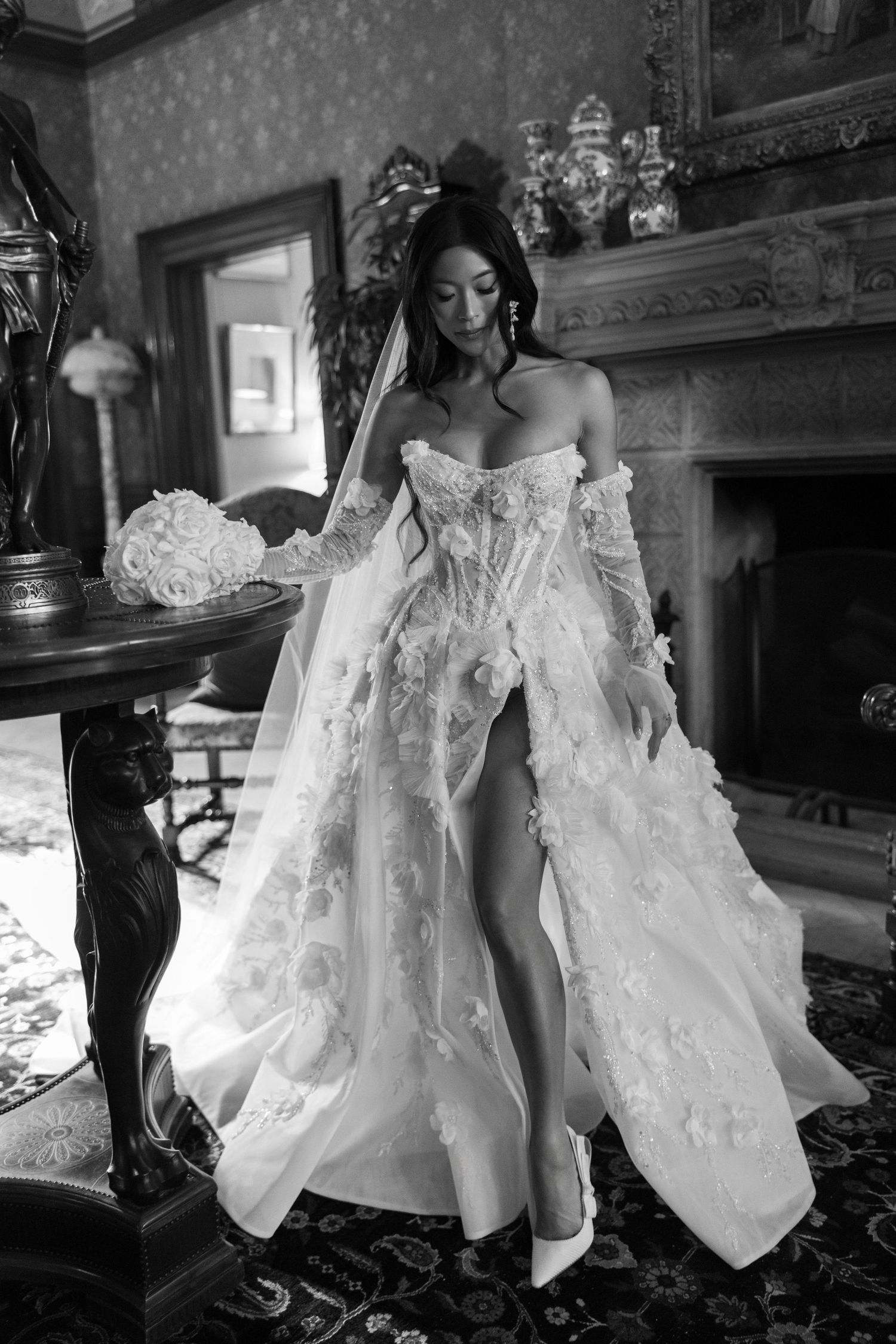 Bride in floral lace off-shoulder wedding gown with thigh-high slit poses in elegant black and white interior.