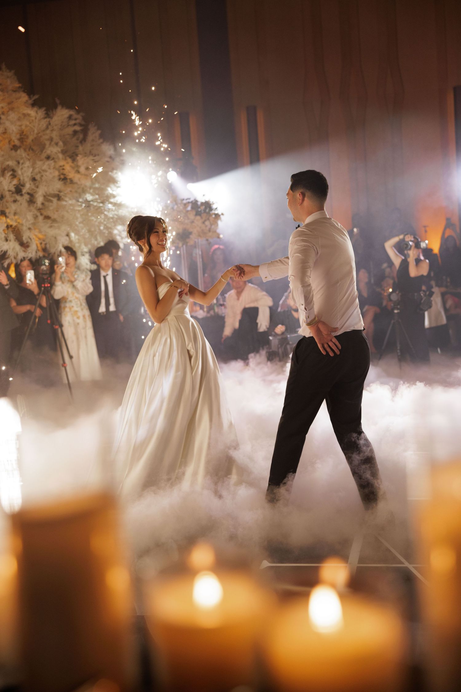 Bride and groom dance on fog-covered floor surrounded by candles and floral arrangements at elegant wedding reception.