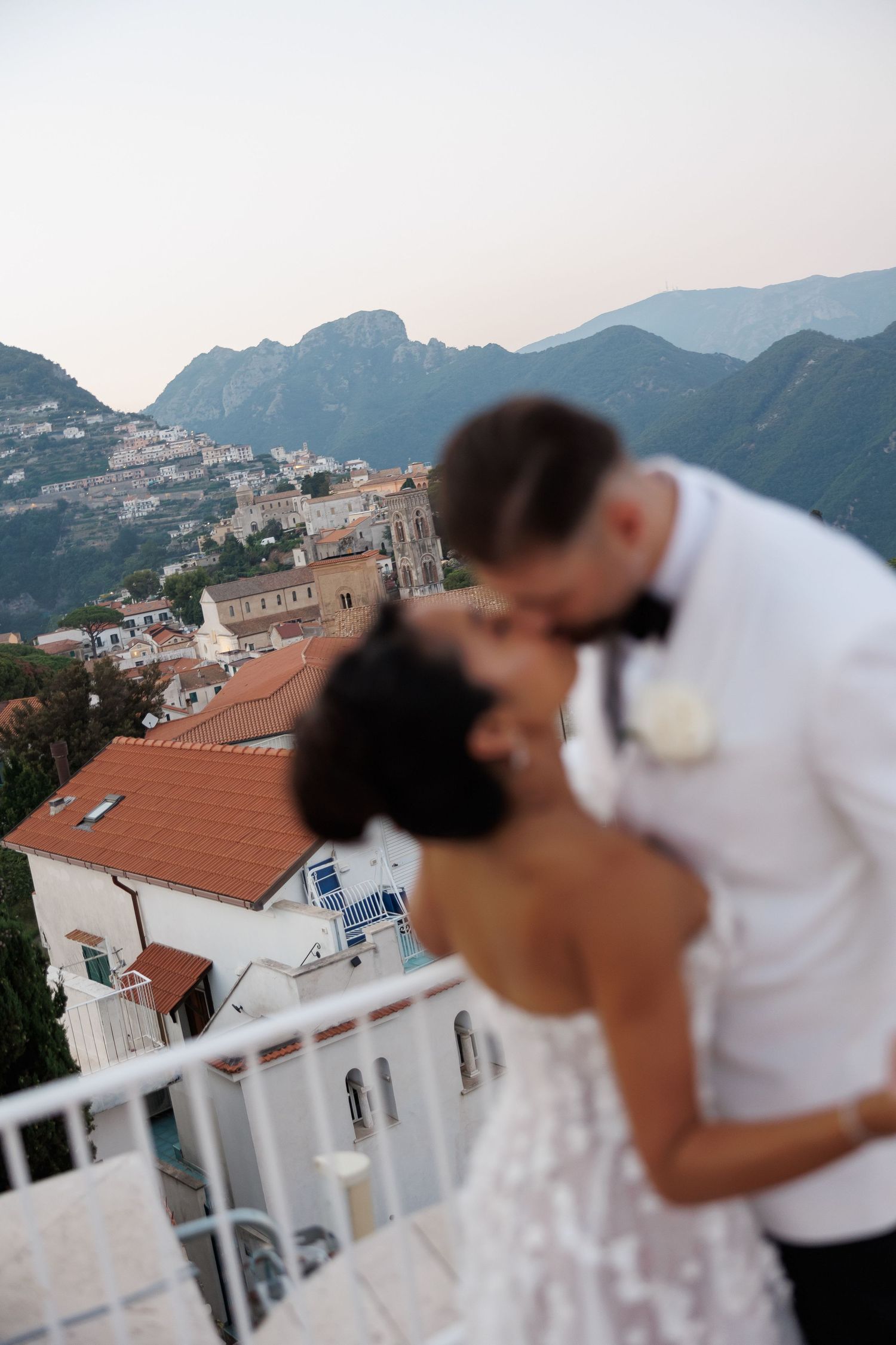Newlyweds share a kiss on a balcony overlooking a scenic Italian coastal mountain village.