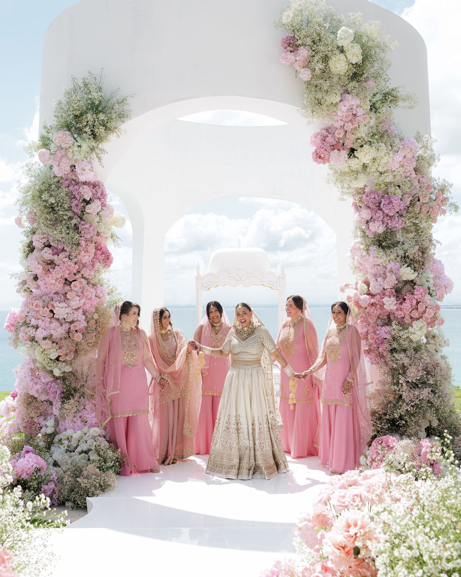 Indian bride with bridesmaids in pink outfits under floral arch with ocean view at destination wedding.