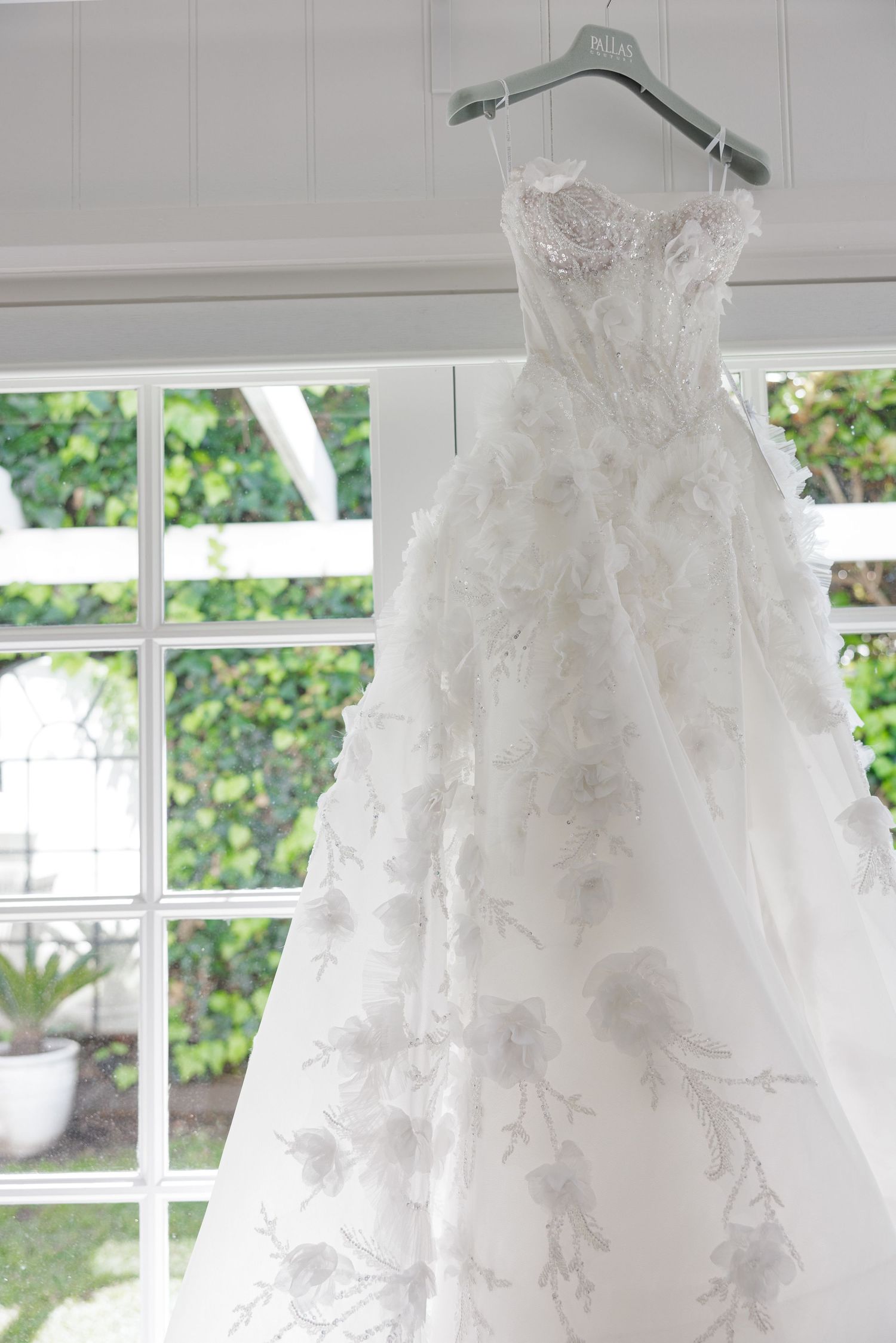 Elegant floral applique ball gown wedding dress hanging on hanger by bright window.