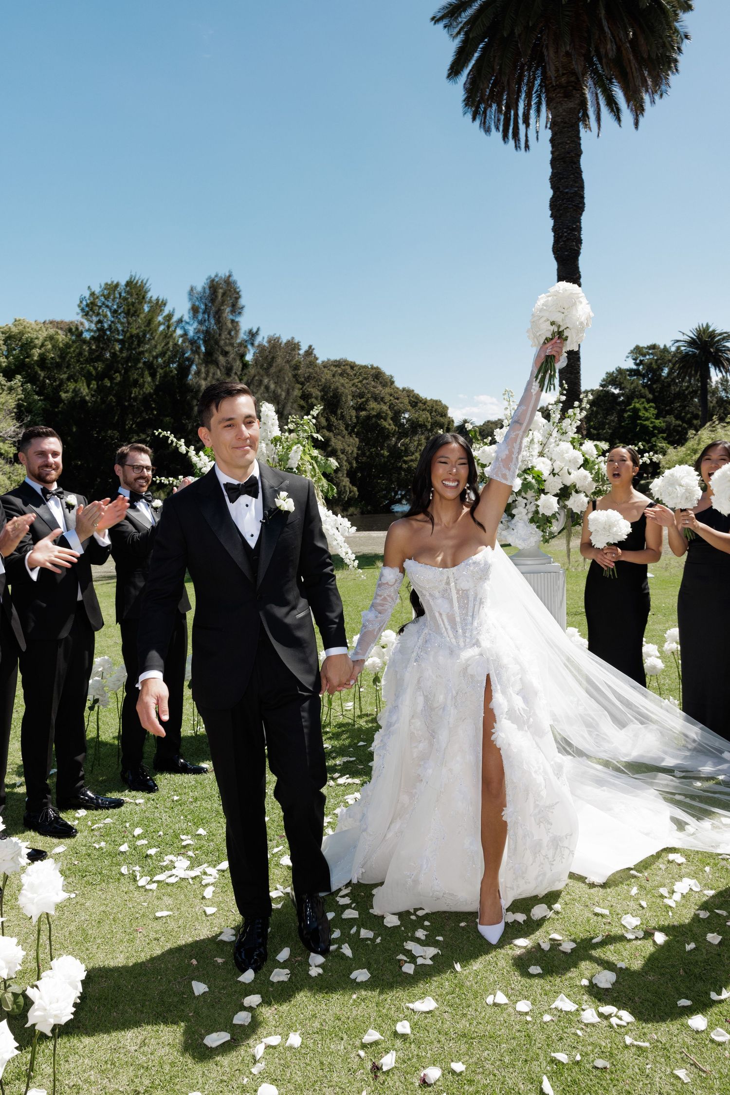 Joyful bride raising bouquet and groom walking hand-in-hand down the aisle at outdoor garden wedding ceremony.