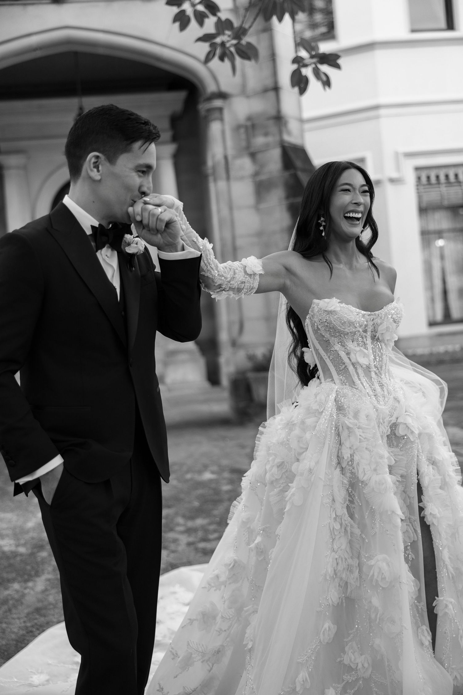 Groom in black tuxedo kisses laughing bride's gloved hand outside historic stone building wedding venue.