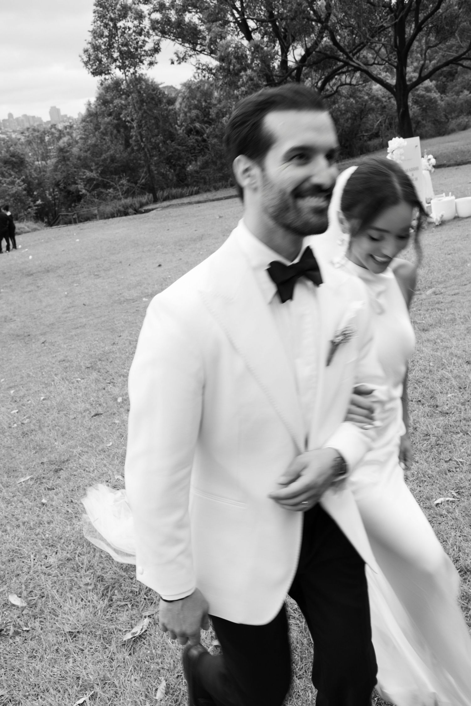 Groom in white tuxedo walks with smiling bride outdoors in black and white photo.