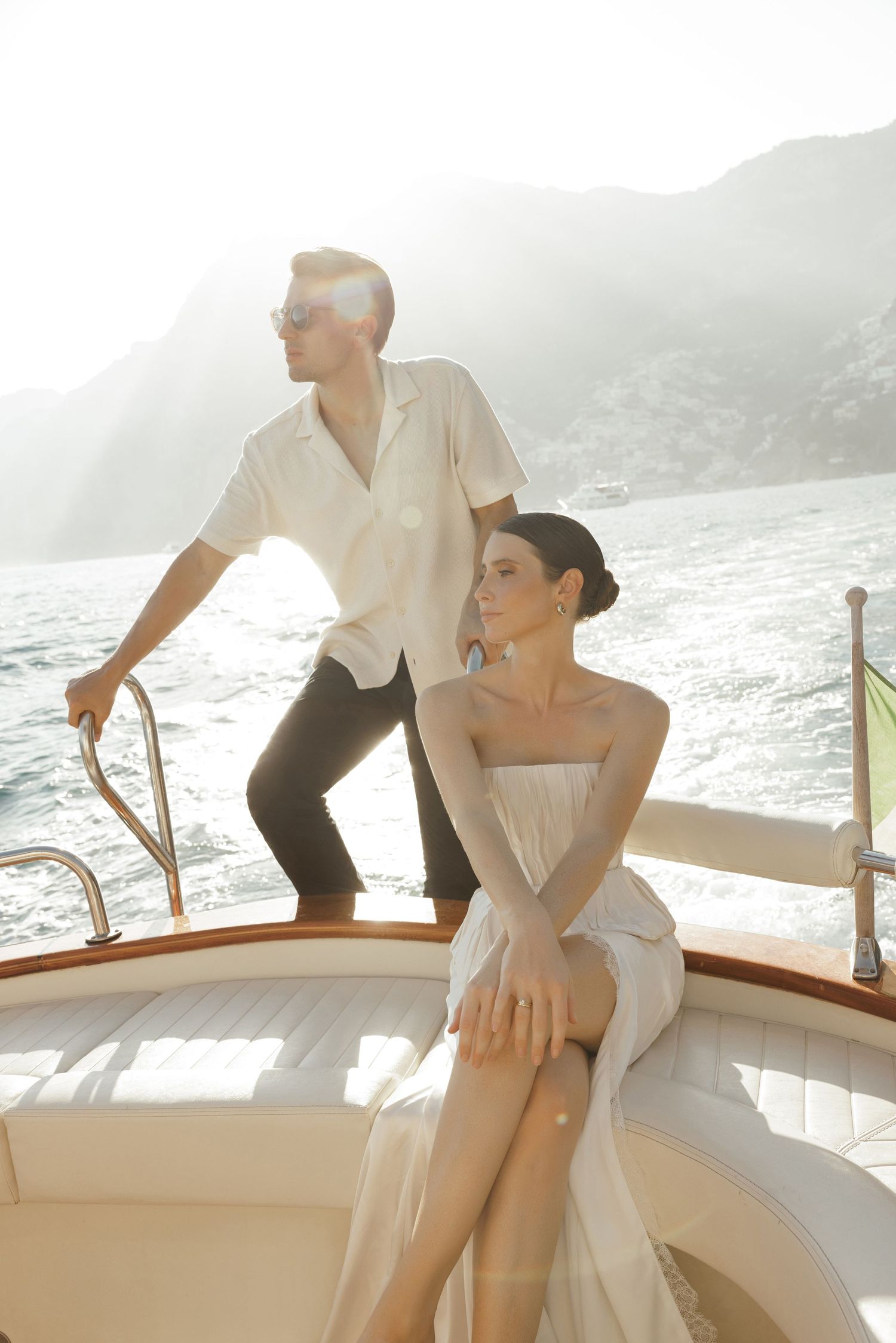 Bride and groom on a luxury yacht with sunlit Mediterranean mountains in background.