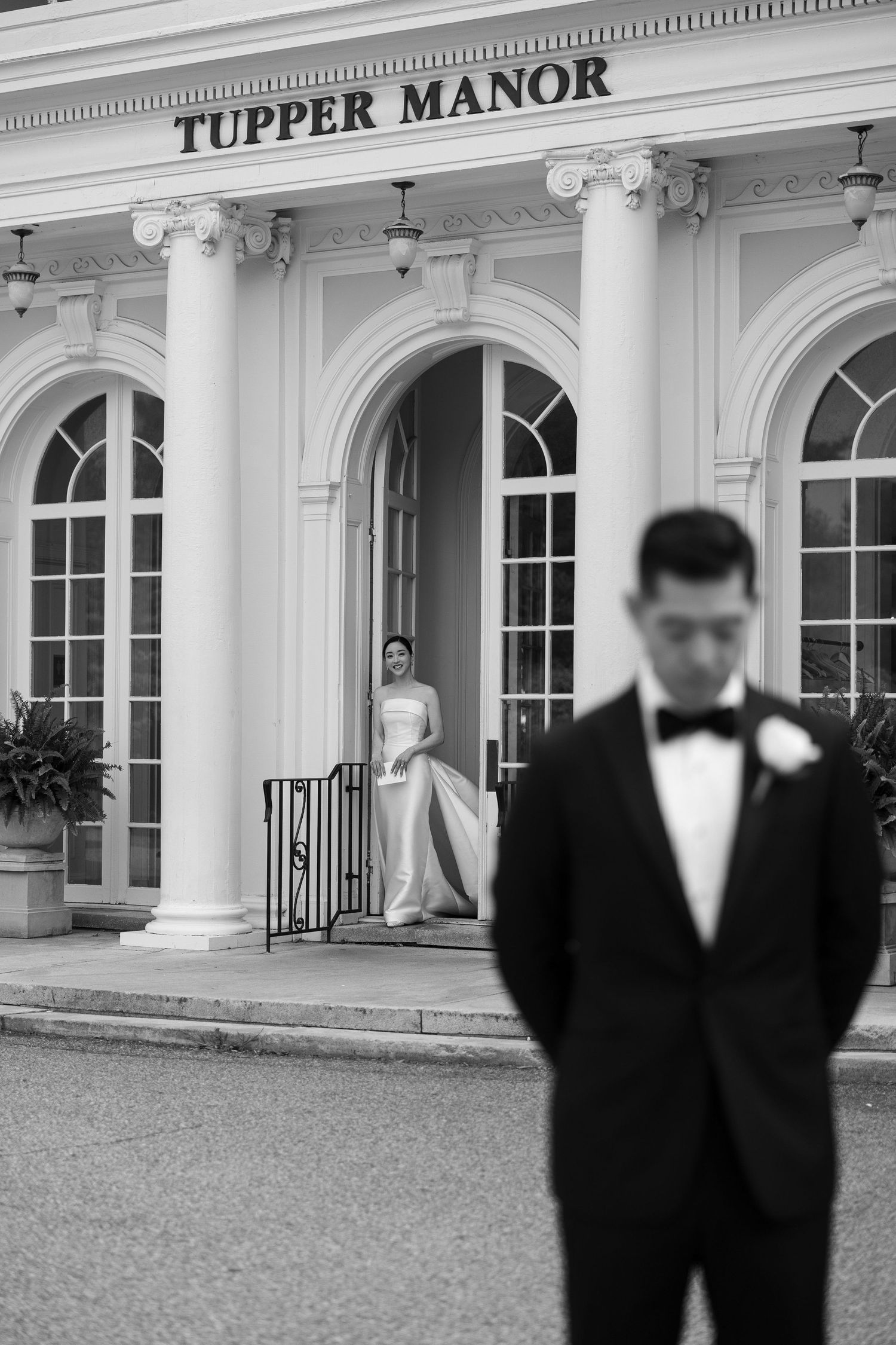 Bride emerges from Tupper Manor as groom stands in foreground in black and white photo.