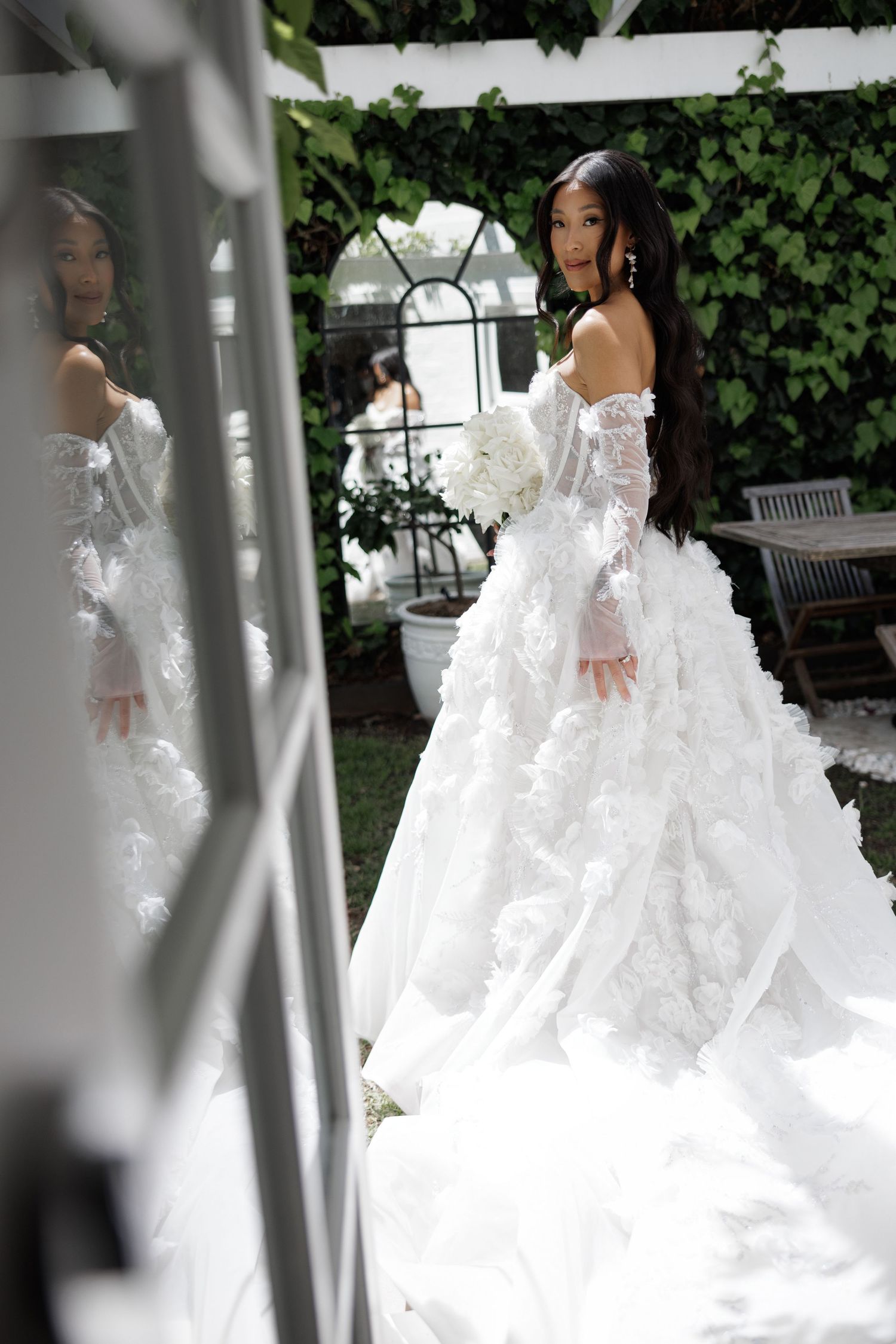 Bride in voluminous off-shoulder floral wedding gown reflected in mirror in garden.