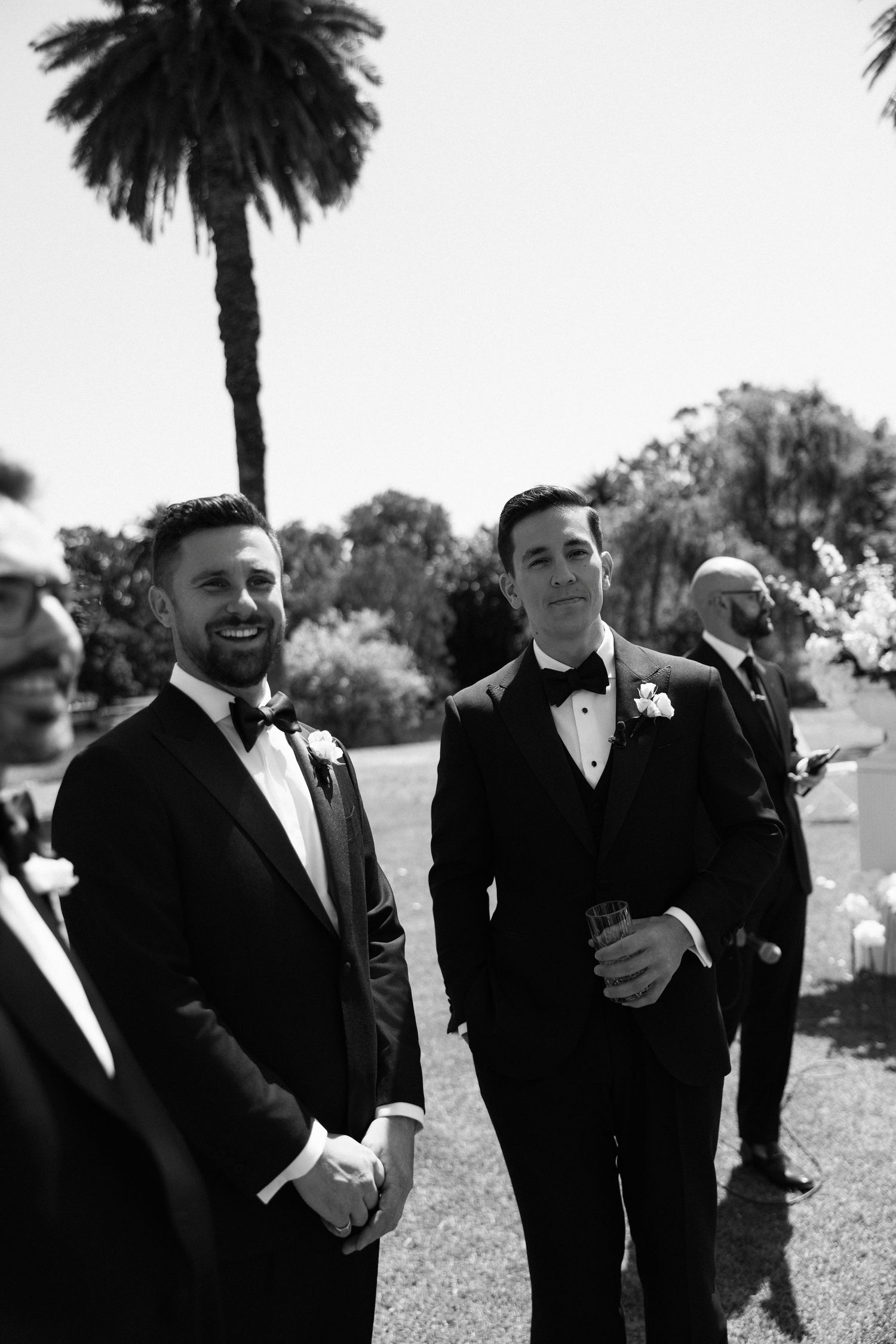 Two groomsmen in black tuxedos with boutonnieres smile outdoors at a wedding ceremony.