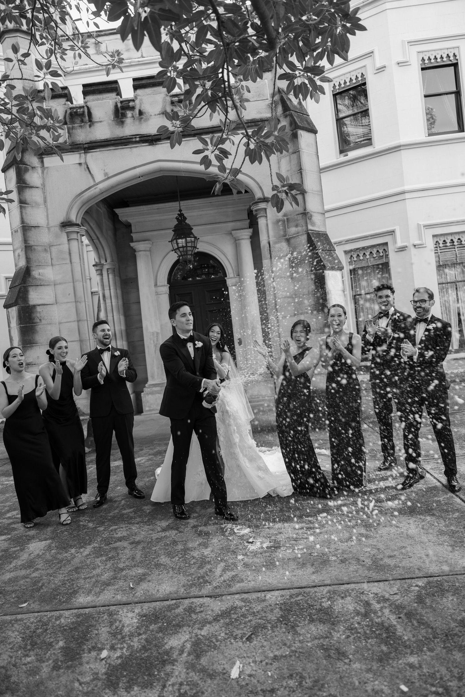Bride and groom celebrate with wedding party as champagne sprays outside elegant stone mansion entrance.