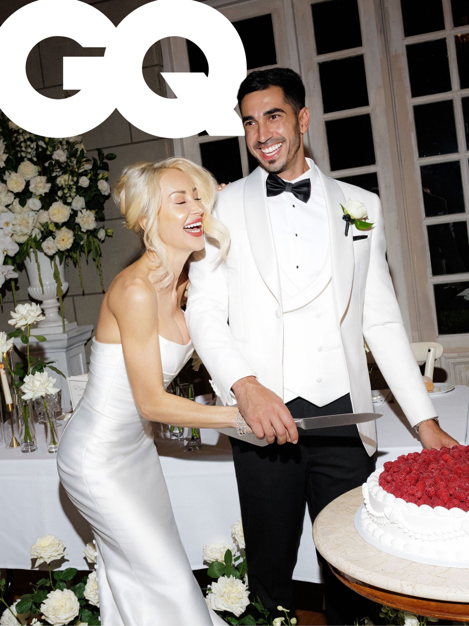 Laughing blonde bride in satin gown and groom in white tuxedo cutting red rose wedding cake at GQ wedding.
