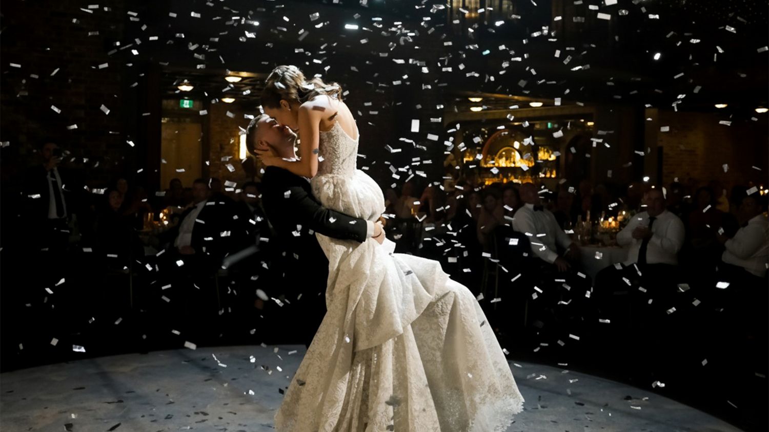 Bride and groom sharing romantic first dance surrounded by falling confetti at wedding reception.