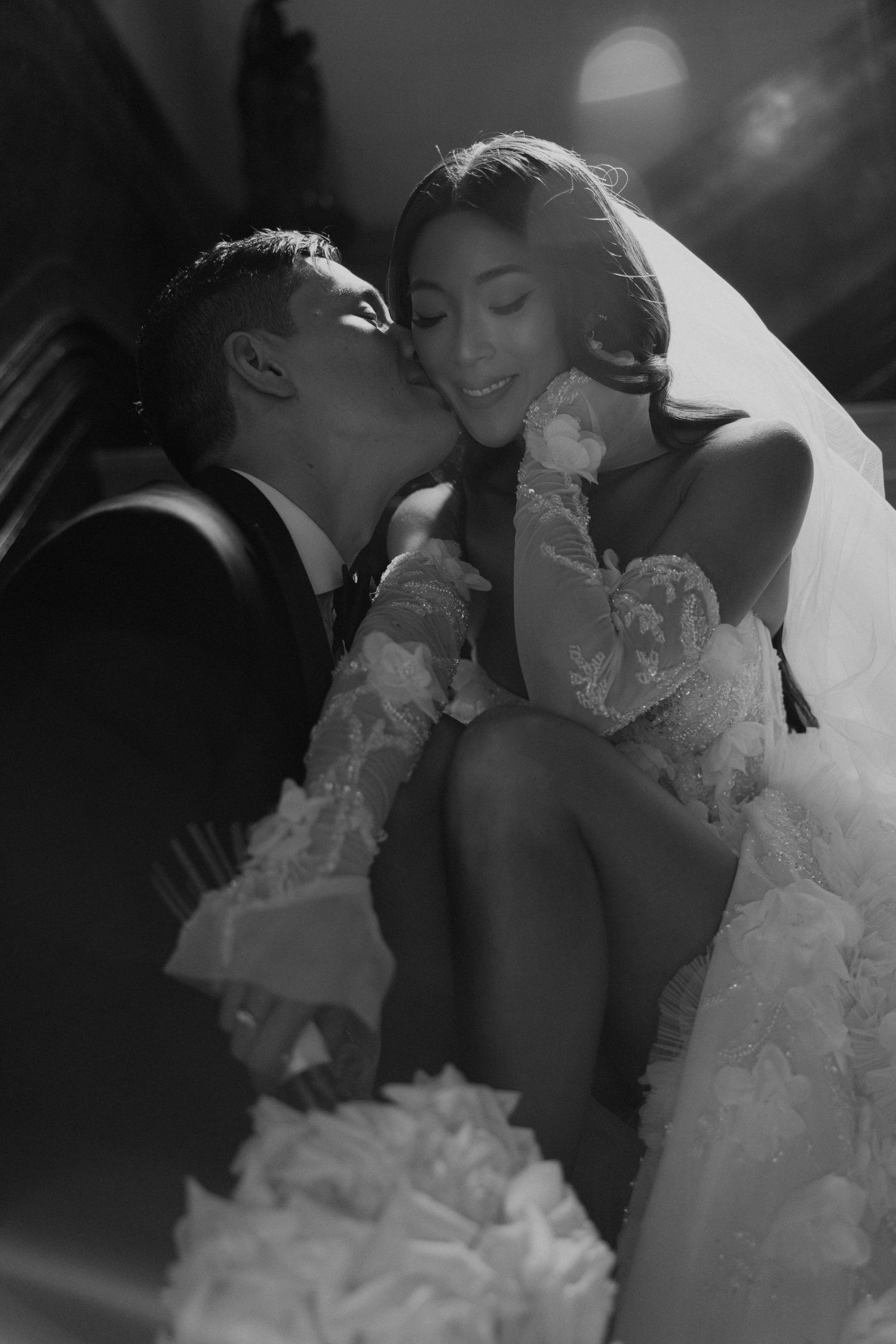 Romantic black and white photo of groom kissing bride on cheek in wedding attire.