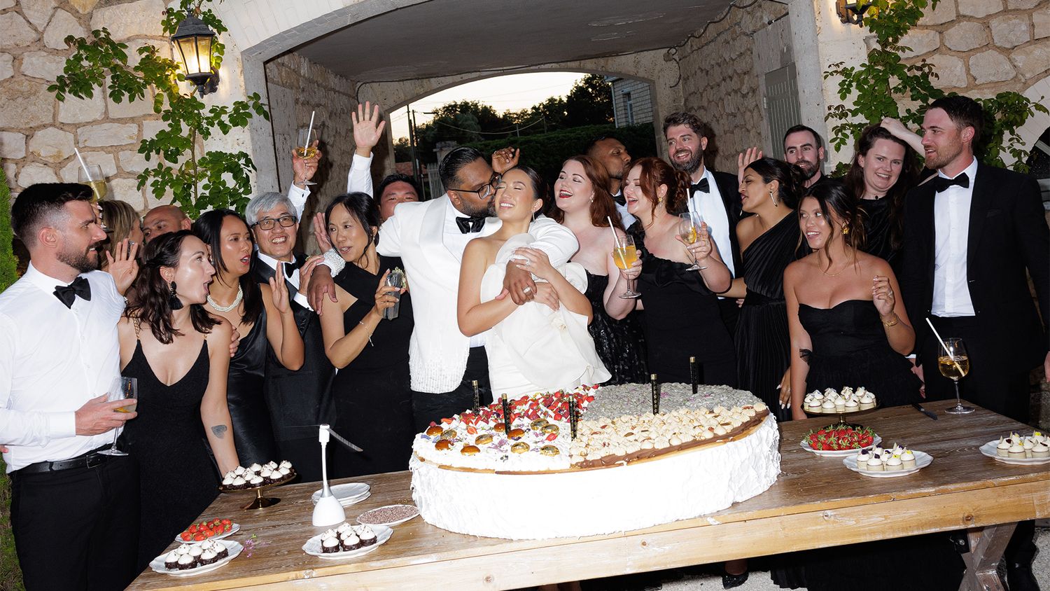 Wedding couple cutting cake surrounded by guests in formal black attire at outdoor reception.