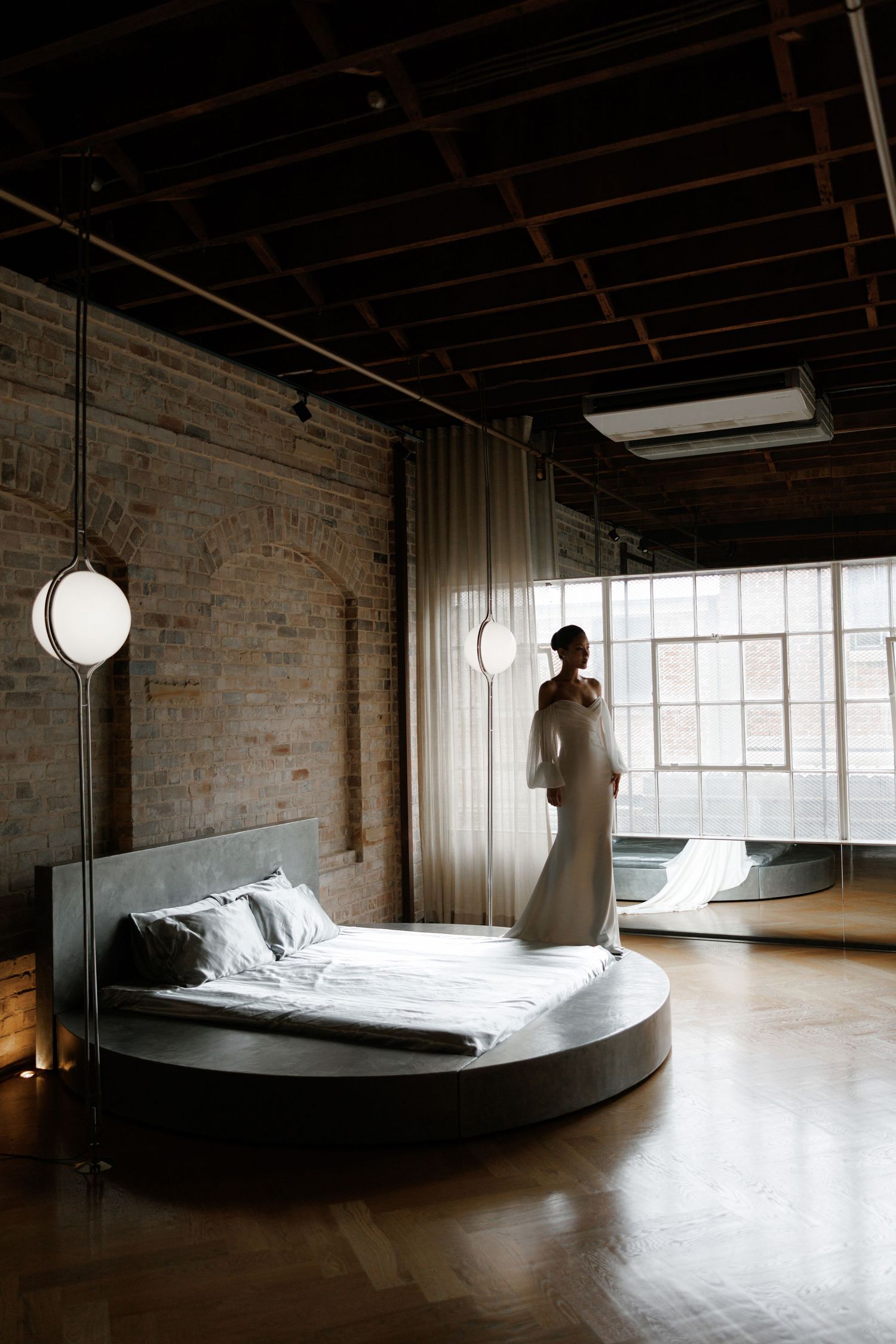 Bride in white gown stands by large industrial loft windows beside a round platform bed.