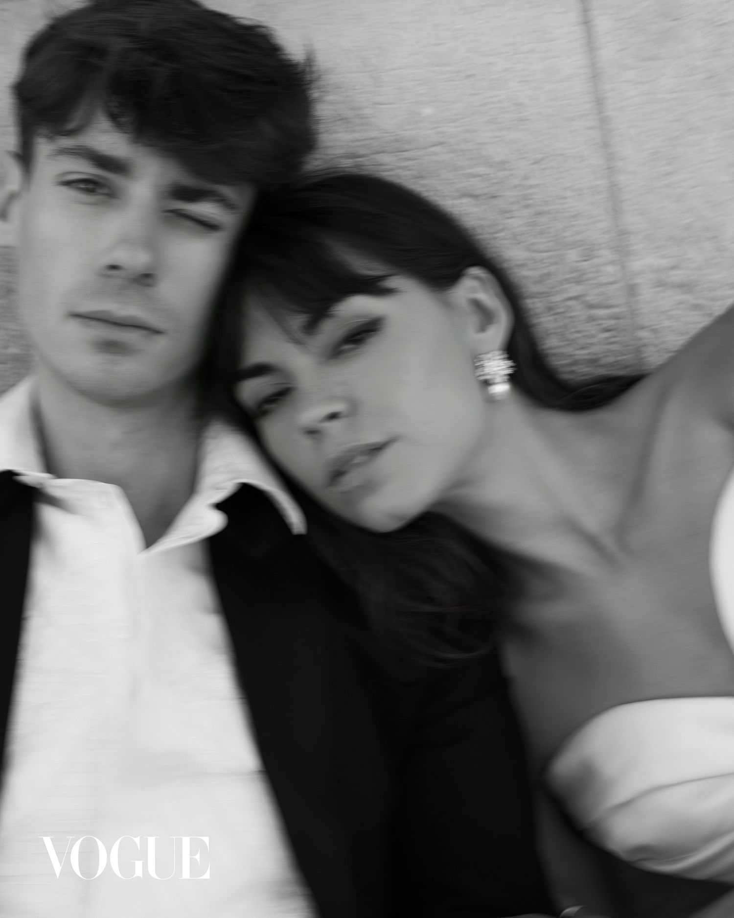 Intimate black and white Vogue portrait of young couple leaning against stone wall together.