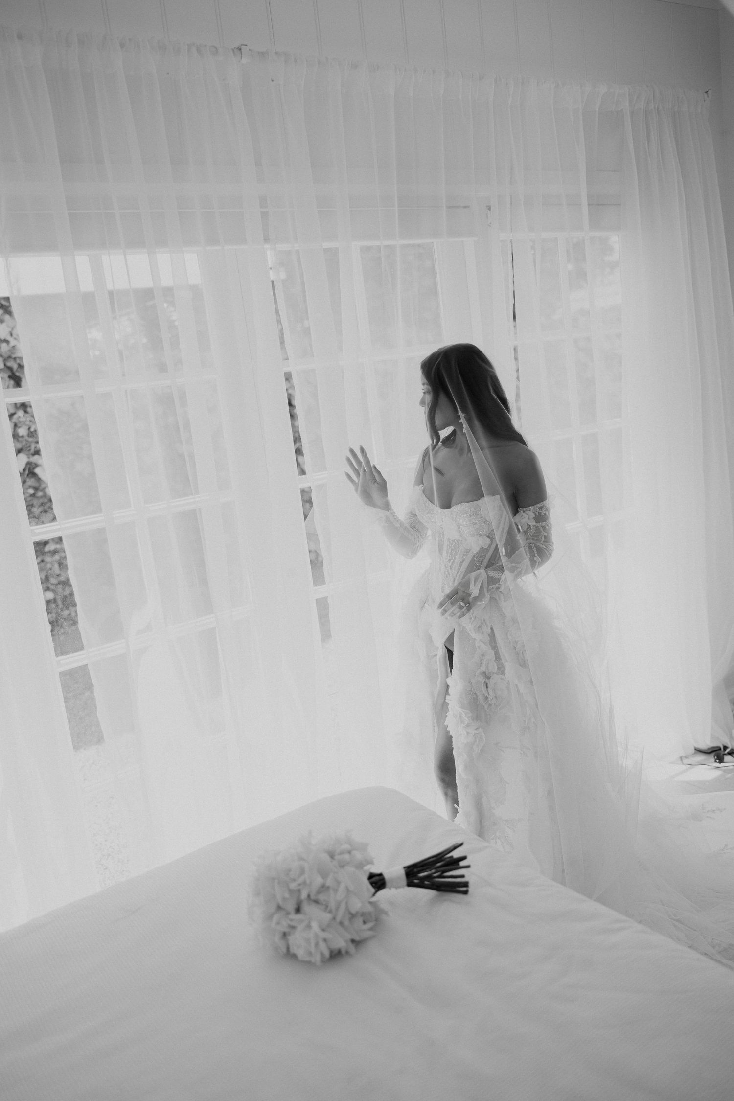 Black and white photo of bride in lace gown standing by sheer curtained window.