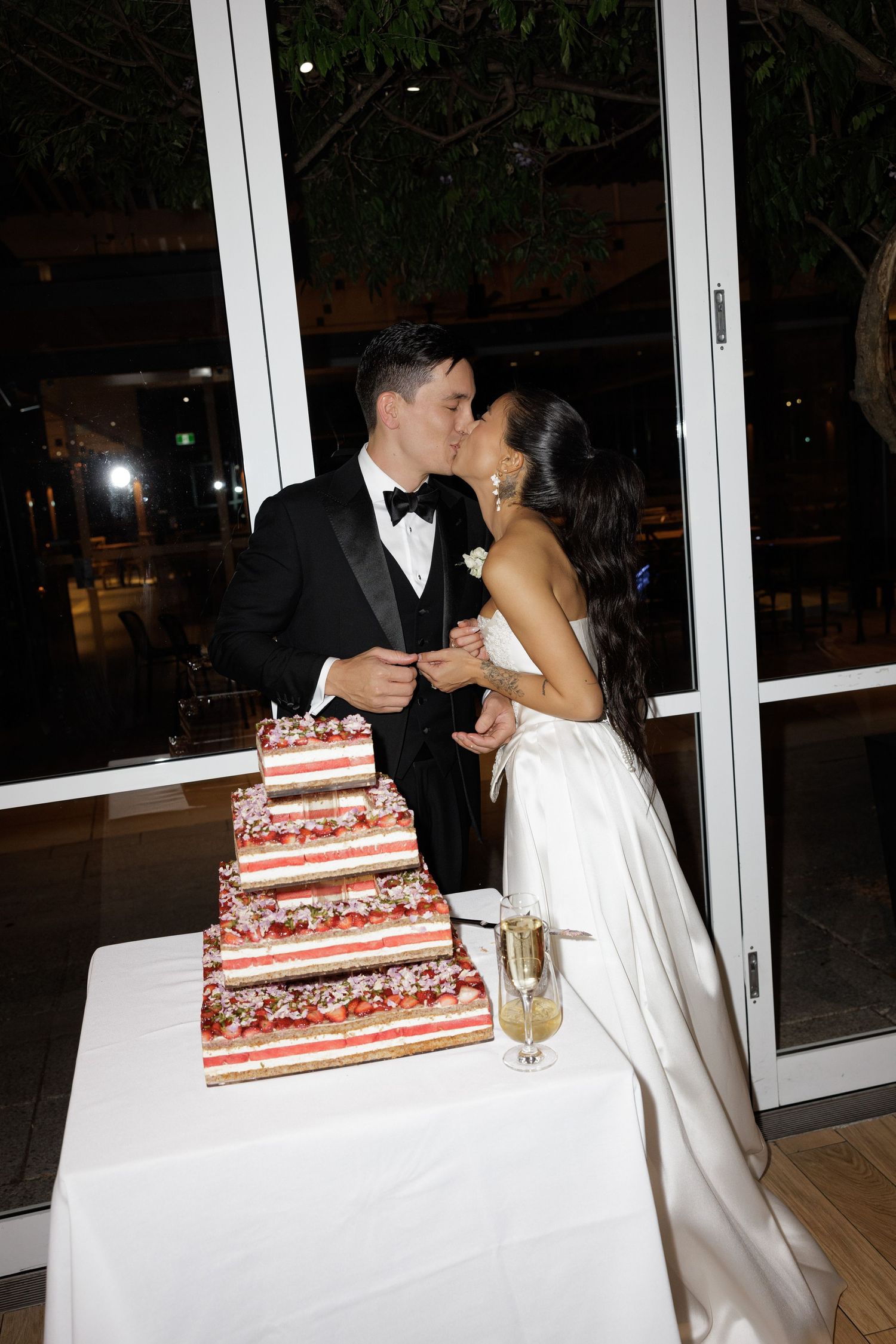 Bride and groom share a kiss while cutting tiered floral wedding cake at reception.